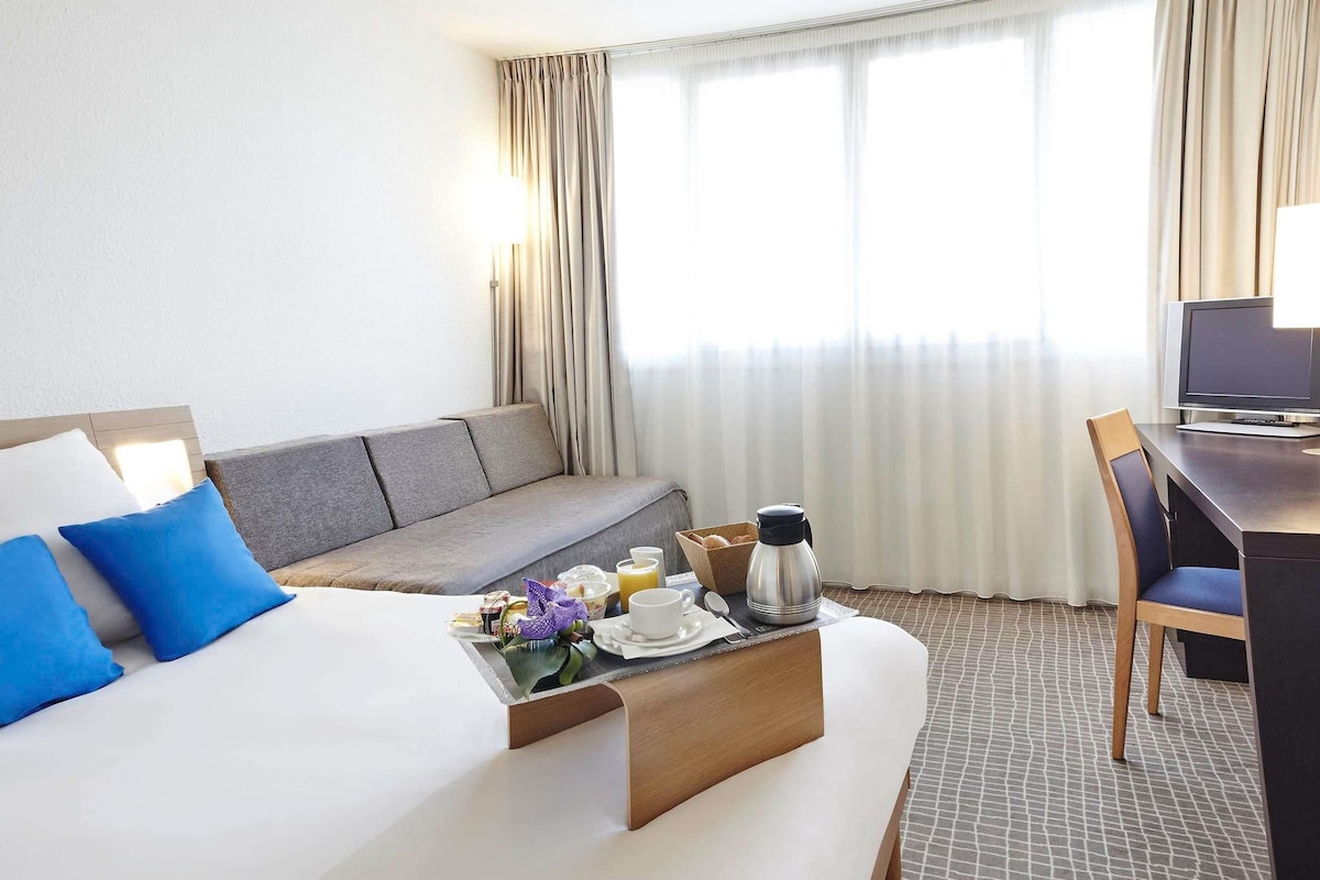 Habitación de hotel con cama, sofá, escritorio con silla, cafetera y bandeja con artículos de desayuno.