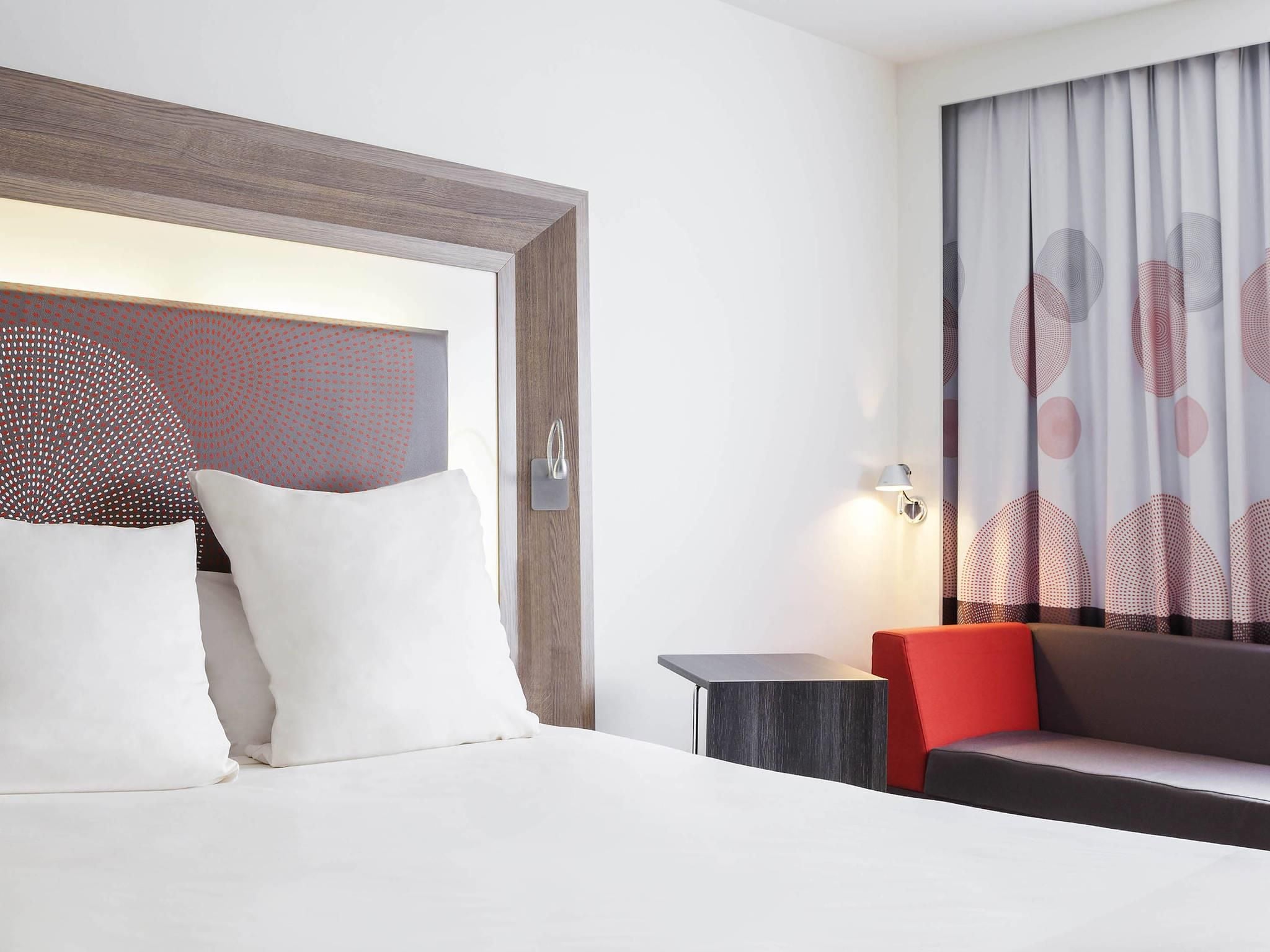 Habitación de hotel con cama, mesita de noche, lámpara, sofá y una pared con diseño geométrico.