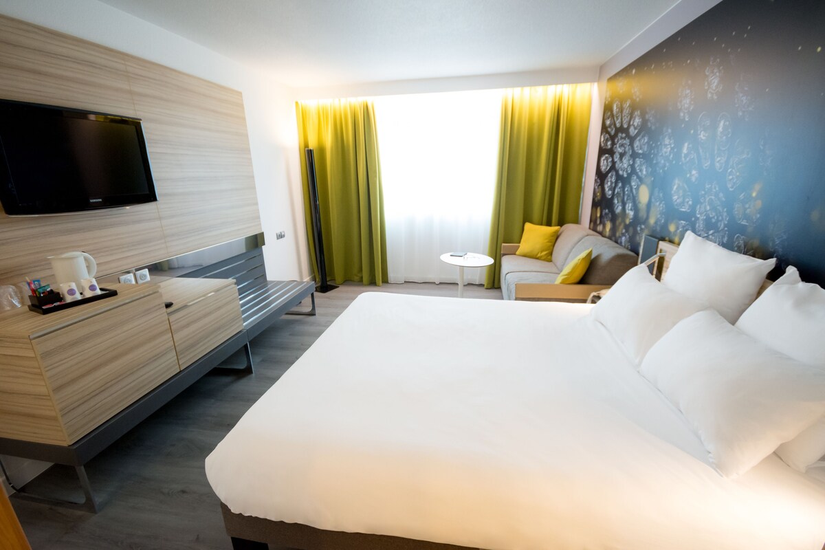A modern hotel room with a large bed, a flat-screen TV mounted on the wall, a small round table, and a sofa with yellow cushions.