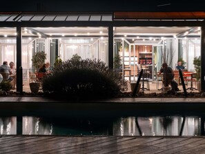 Un área de comedor exterior moderna con paredes de vidrio, una piscina y plantas en macetas.