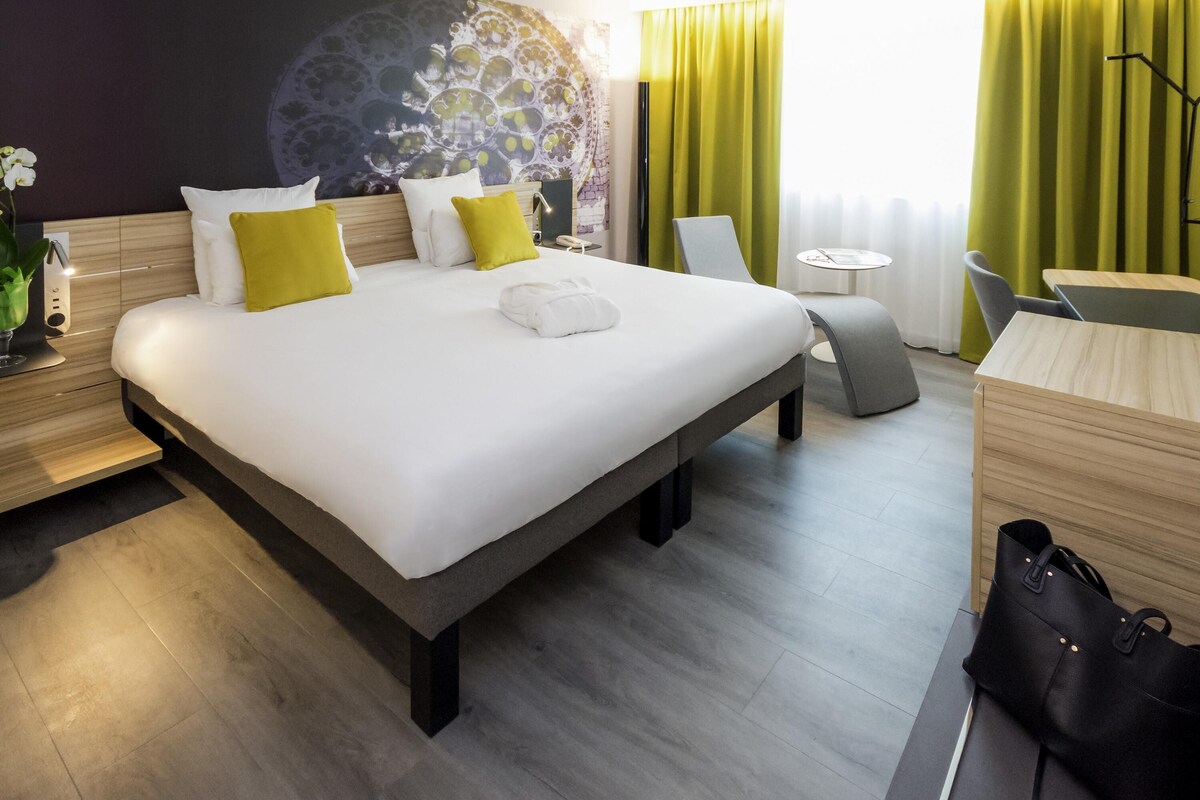 A modern hotel room with a large bed, a desk, a chair, a small table, and a view of the city through the window.