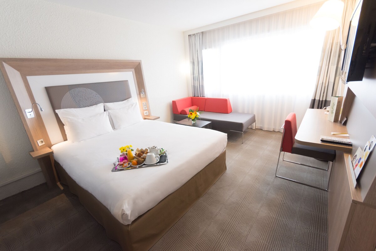 A hotel room with a large bed, a desk, a red sofa, and a TV.