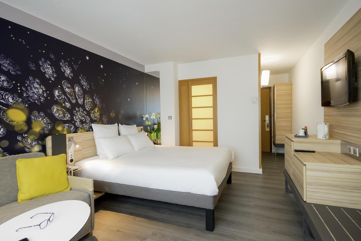 Una habitación de hotel moderna con cama, mesita de noche, televisión y una pared con un mural decorativo.