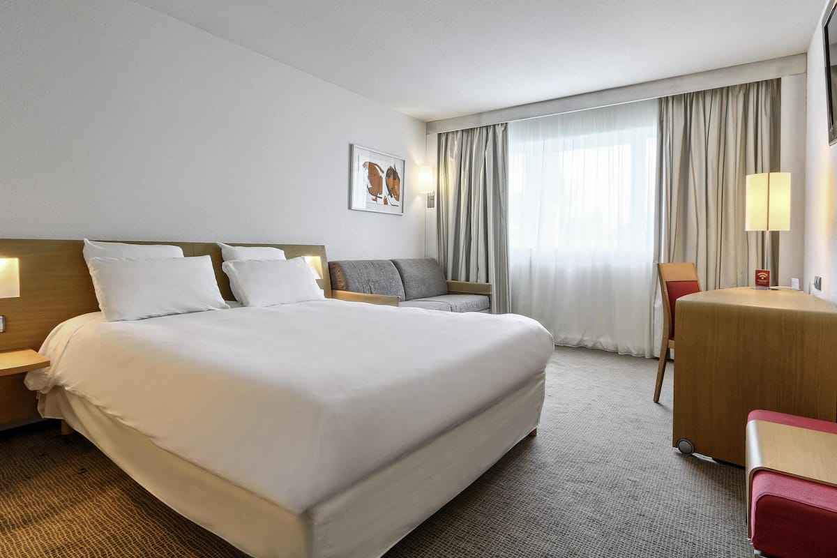 Ein Hotelzimmer mit einem großen Bett, einem Schreibtisch, einem Stuhl, einer Lampe und einem Vorhangfenster.