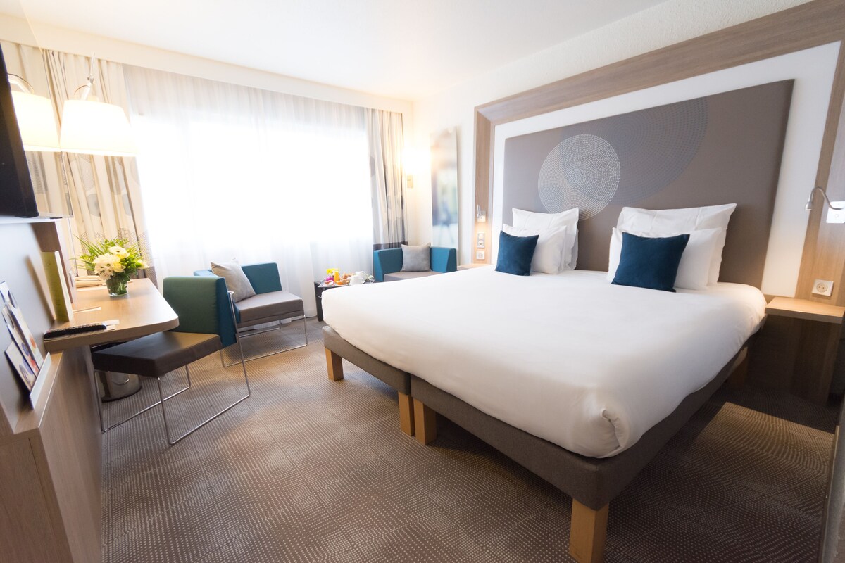 A hotel room with a large bed, a desk with a chair, a TV, and a window with curtains.