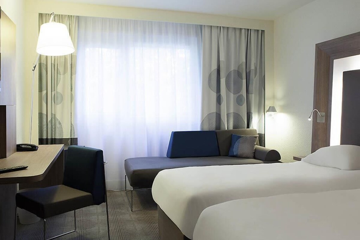 A hotel room with a bed, a desk, a chair, and a large window with curtains.