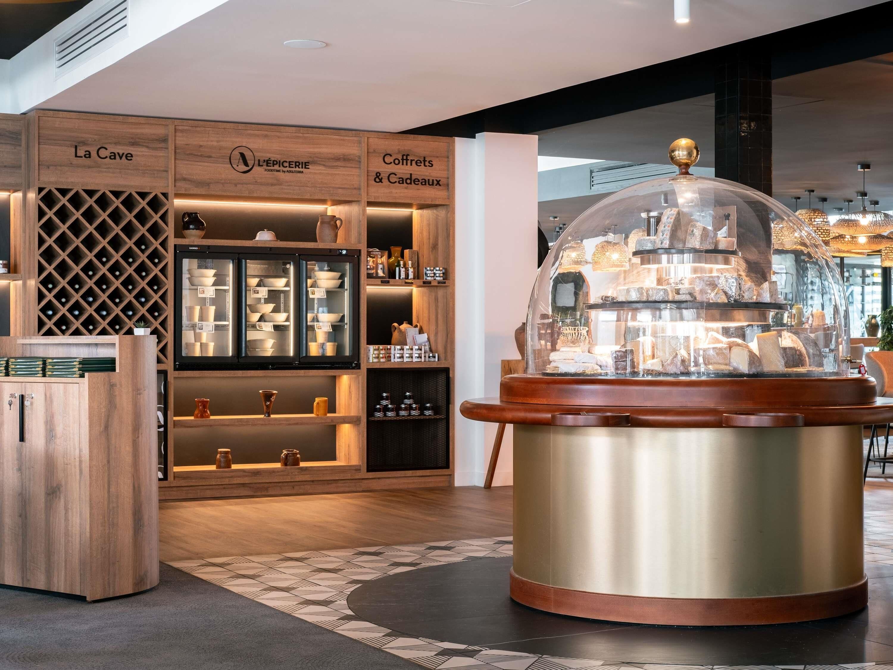 A modern interior with a wine cooler, wooden shelves labeled "La Cave," "L'EPIERIE," and "Coffrets & Cadeaux," and a central display case with a dome.
