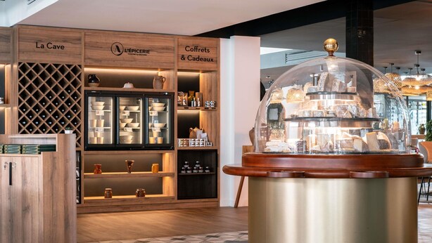 A modern interior with a wine cooler, wooden shelves labeled "La Cave," "L'EPIERIE," and "Coffrets & Cadeaux," and a central display case with a dome. - Novotel Chartres (Chartres)