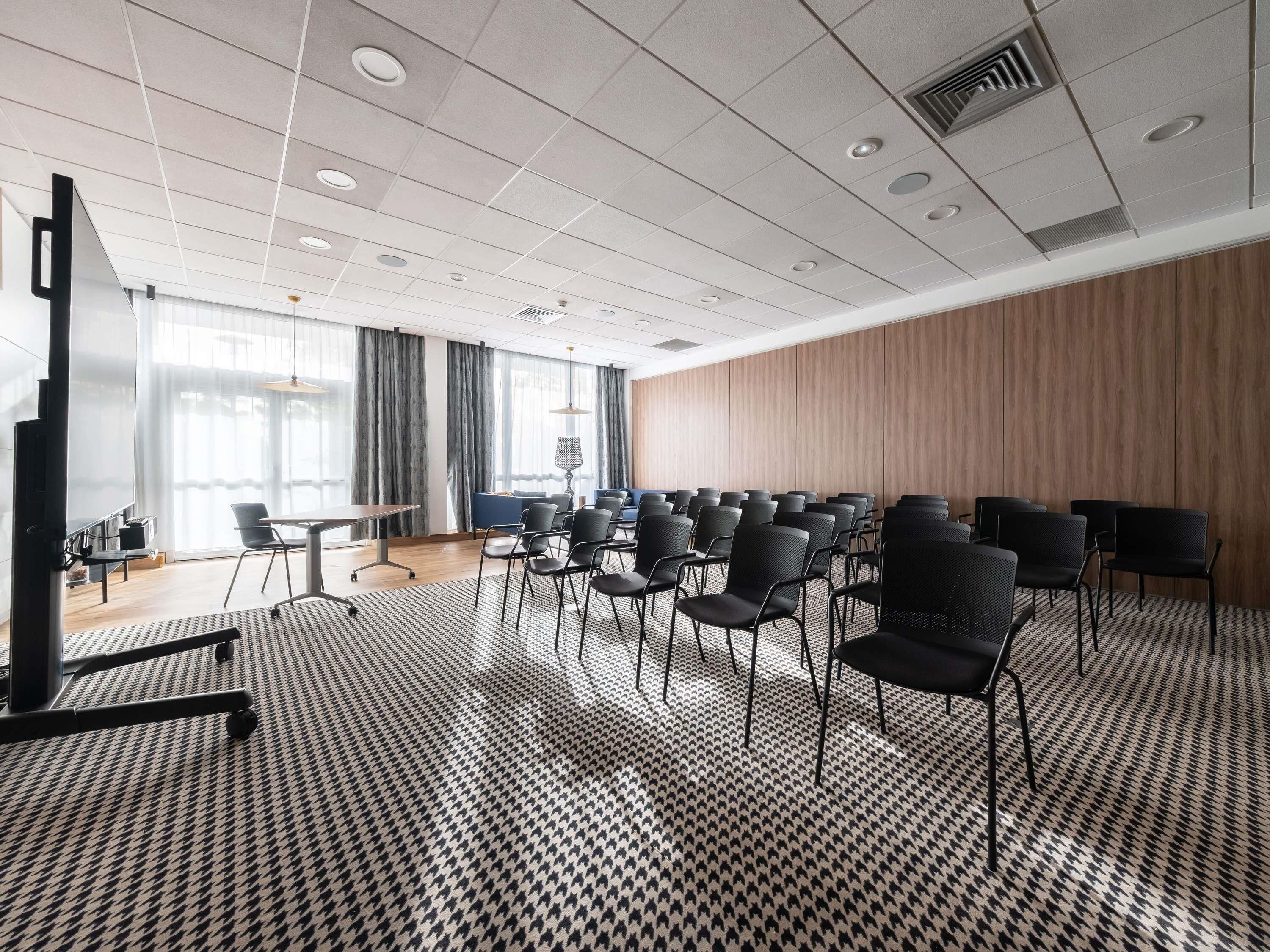 Un salón de conferencias con alfombra a cuadros de galgo, filas de sillas, una pantalla grande y un panel de pared de madera.