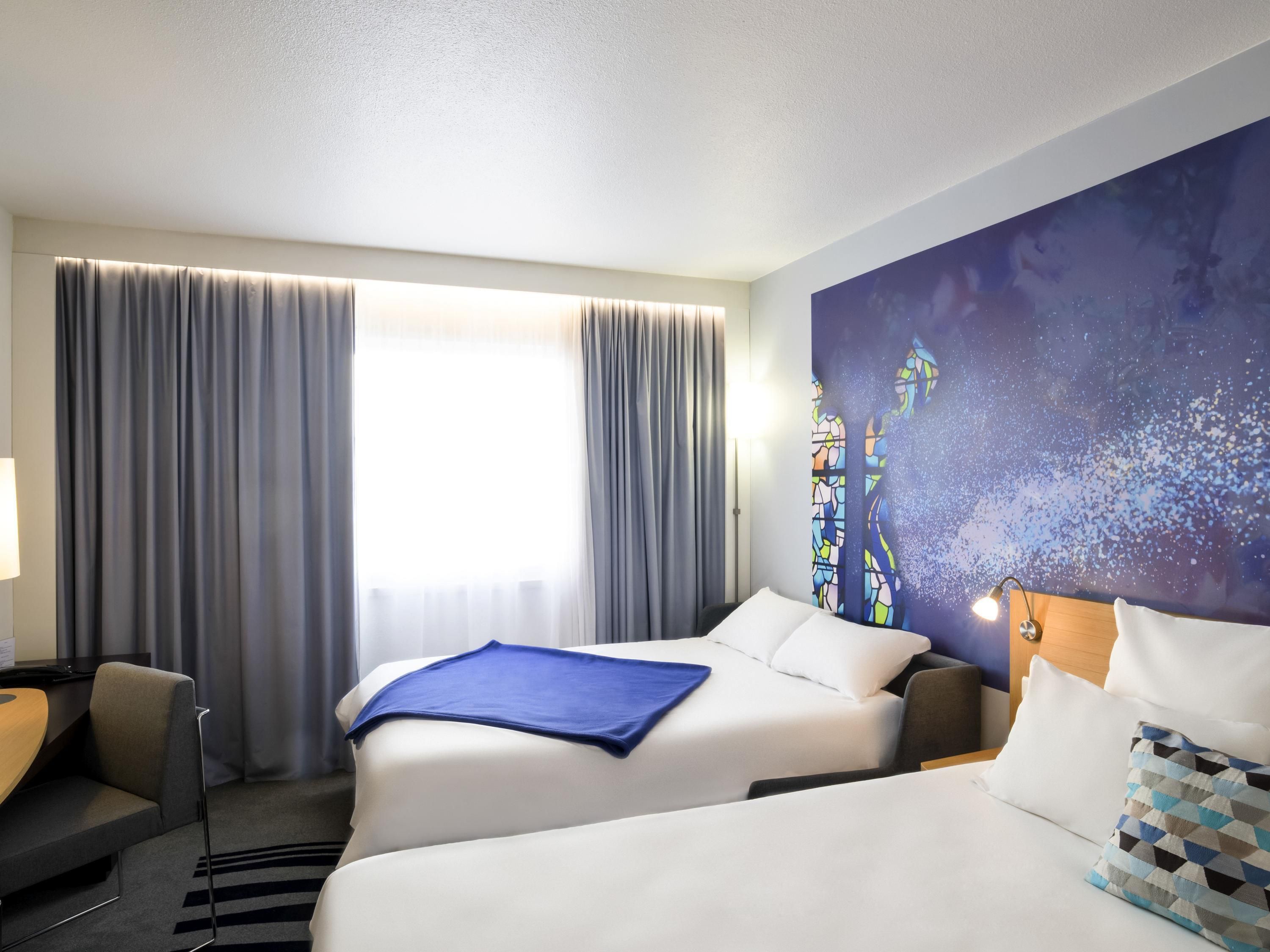 A hotel room with two beds, a desk, a chair, a large window with curtains, and a wall mural.