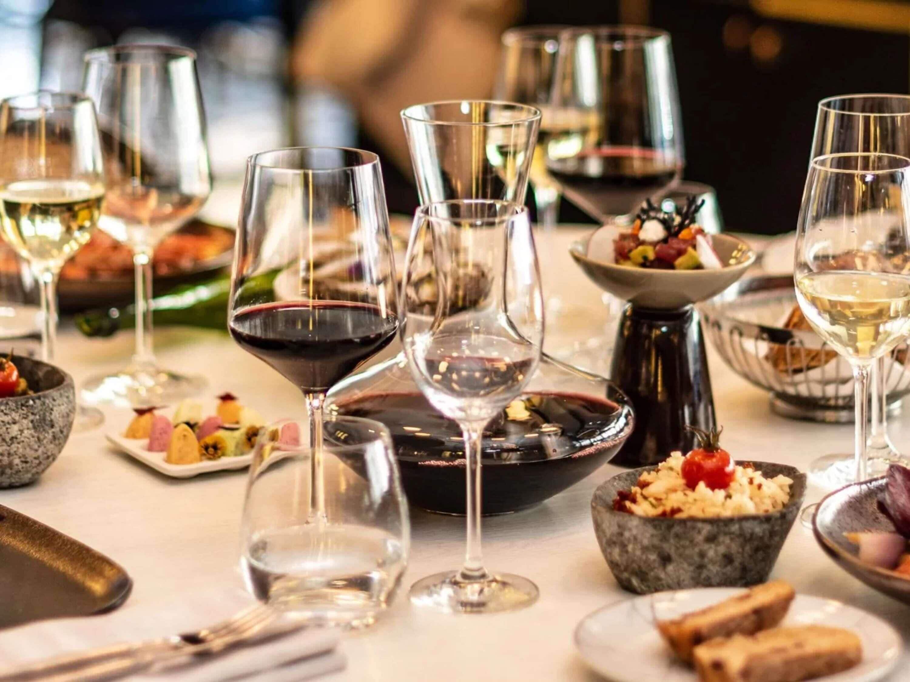 Una mesa de comedor con varias copas de vino, fuentes de comida y una jarra de metal.