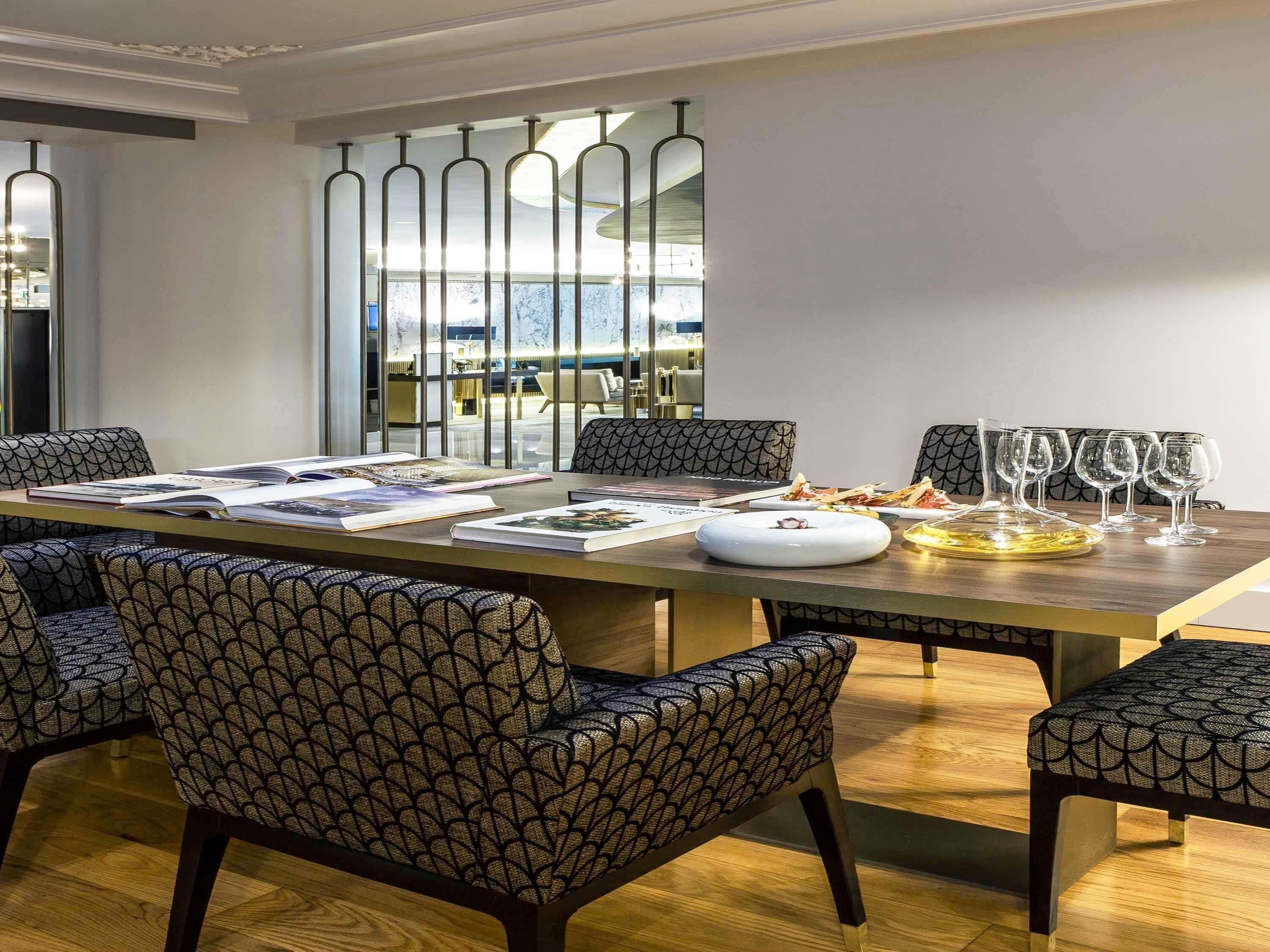 A modern dining area with a long table, patterned chairs, and a view of the city through large windows.