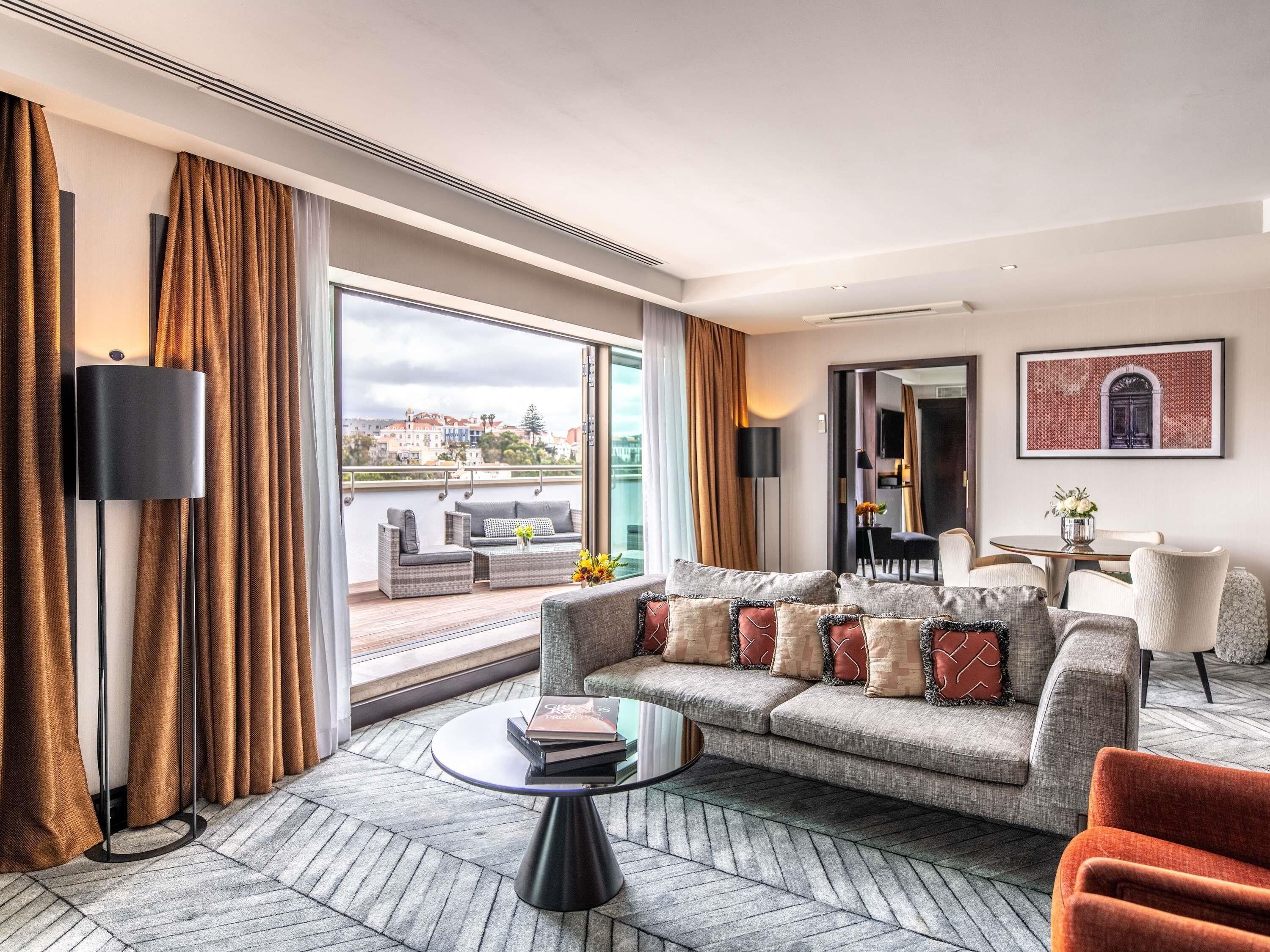 Una habitación de hotel moderna con un balcón grande, un conjunto de sofás, un comedor y vistas a la ciudad.