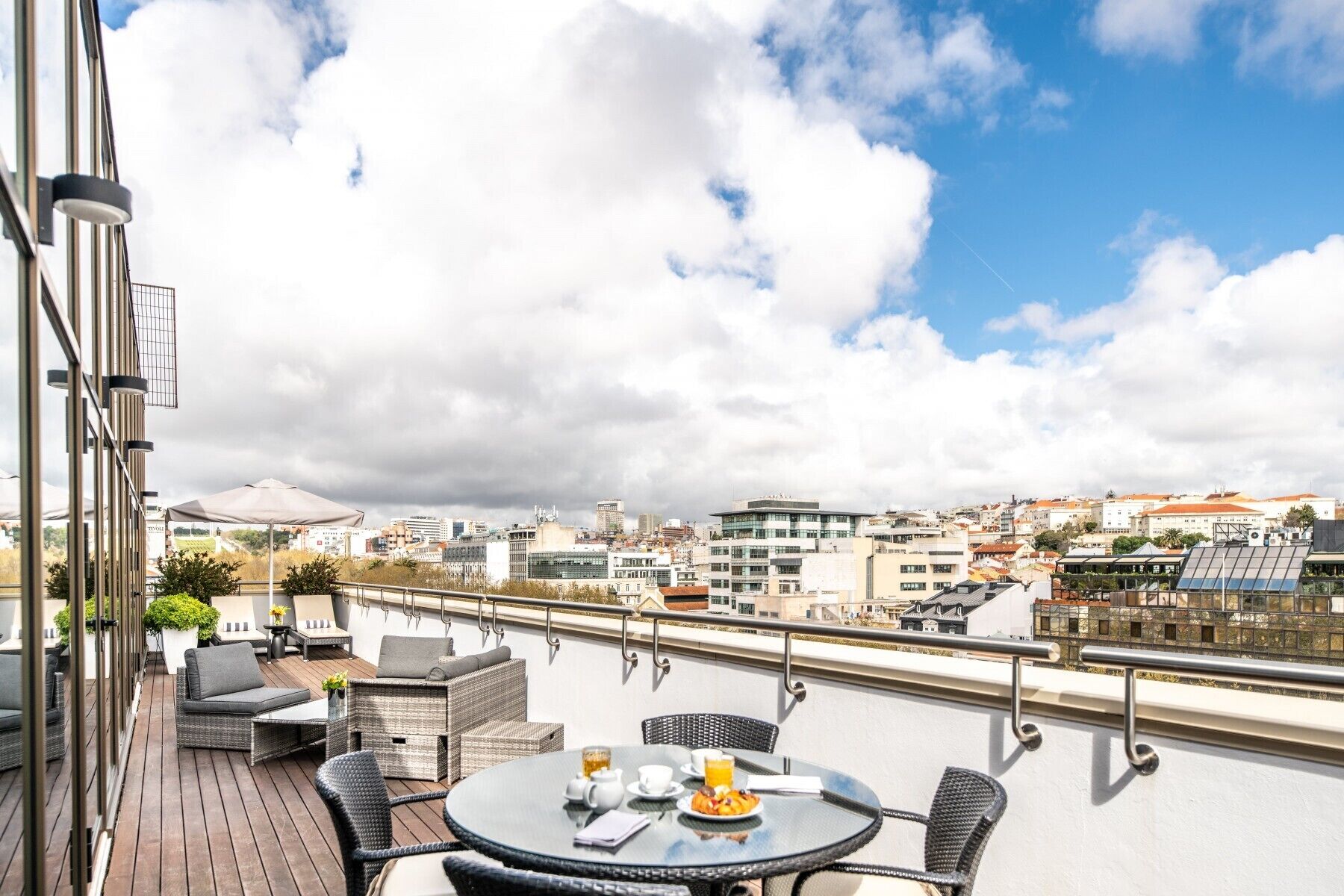 Una terraza en la azotea con mobiliario exterior, una mesa con desayuno y vistas a la ciudad.