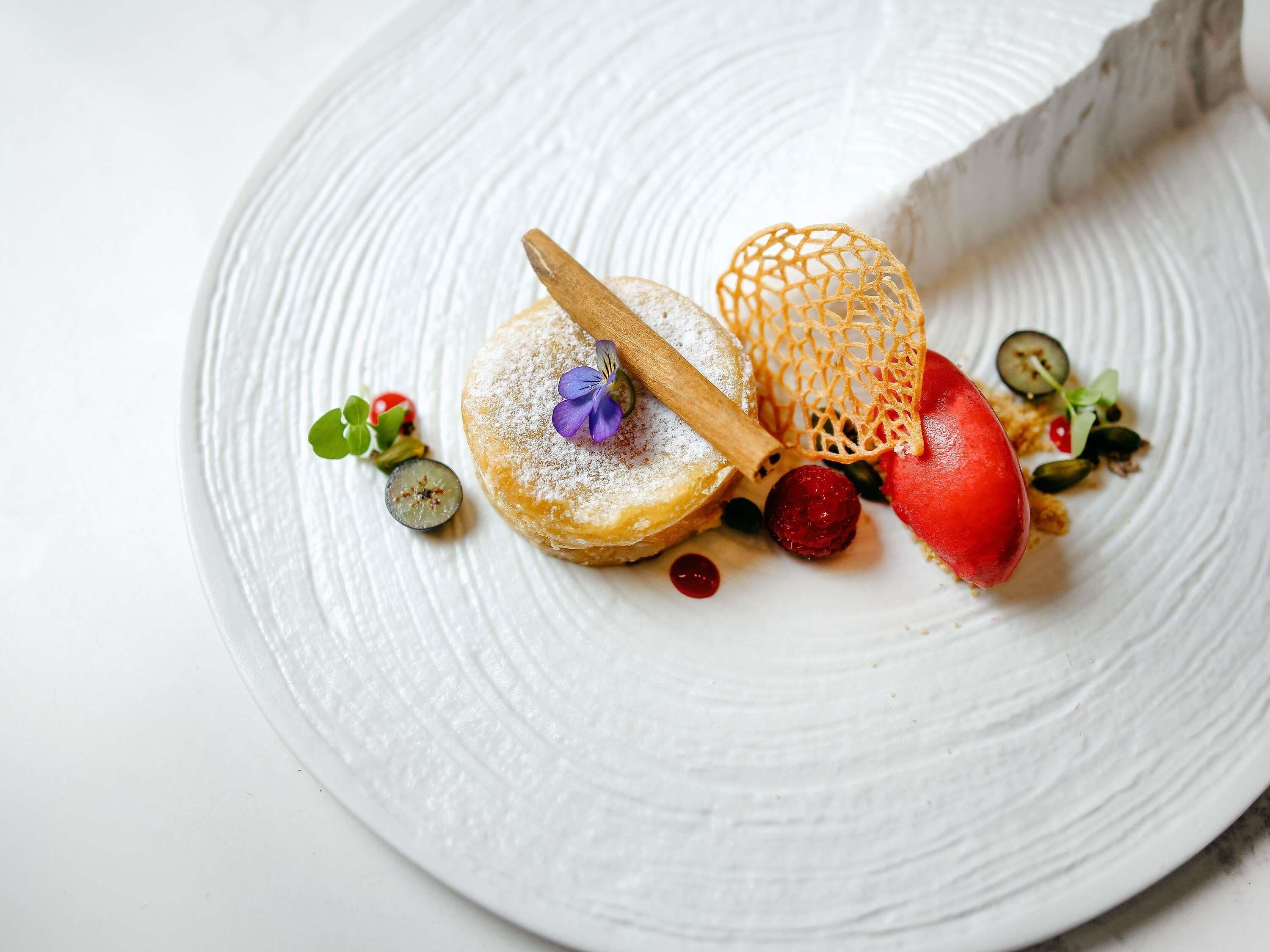 A dessert plate with a pastry, a wooden stick, a netted item, and various garnishes.