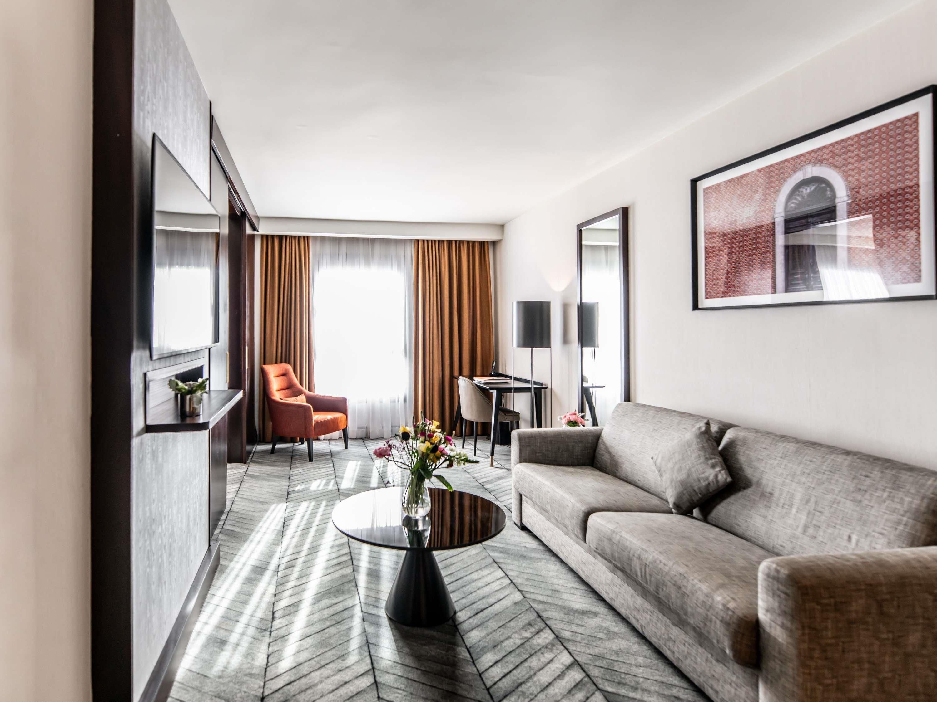 A modern hotel room with a sofa, armchair, desk, and a large window with curtains.