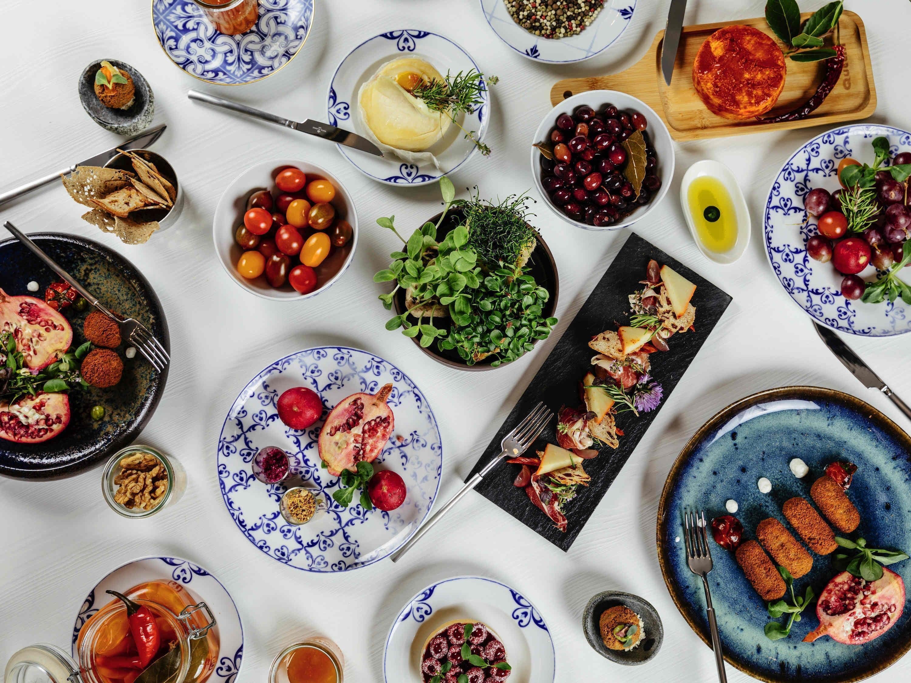 A variety of dishes on a table, including a pomegranate, cherry tomatoes, and a bowl of olives.