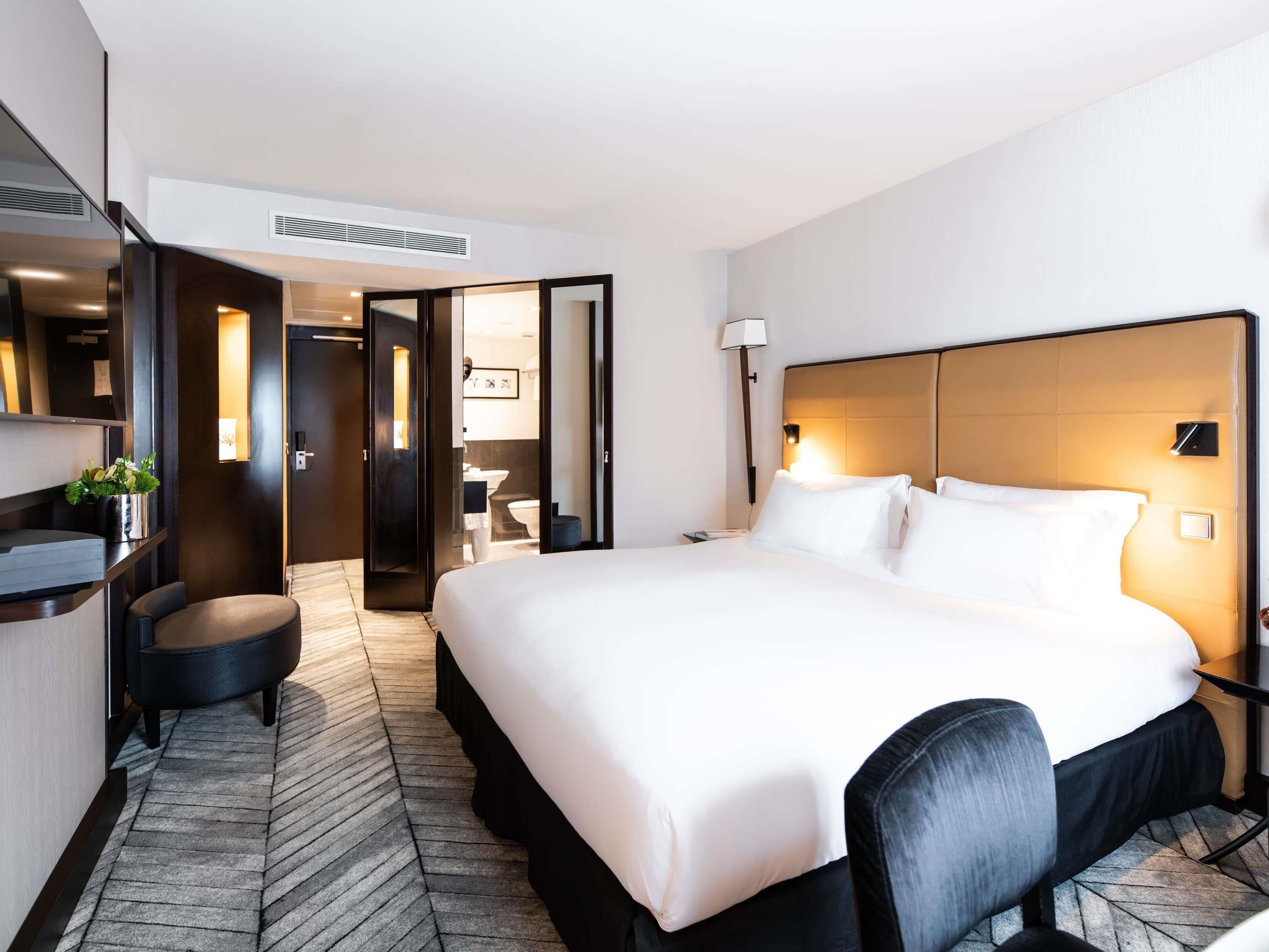 A hotel room with a large bed, a desk with a chair, a mirror, and a bathroom visible through an open door.