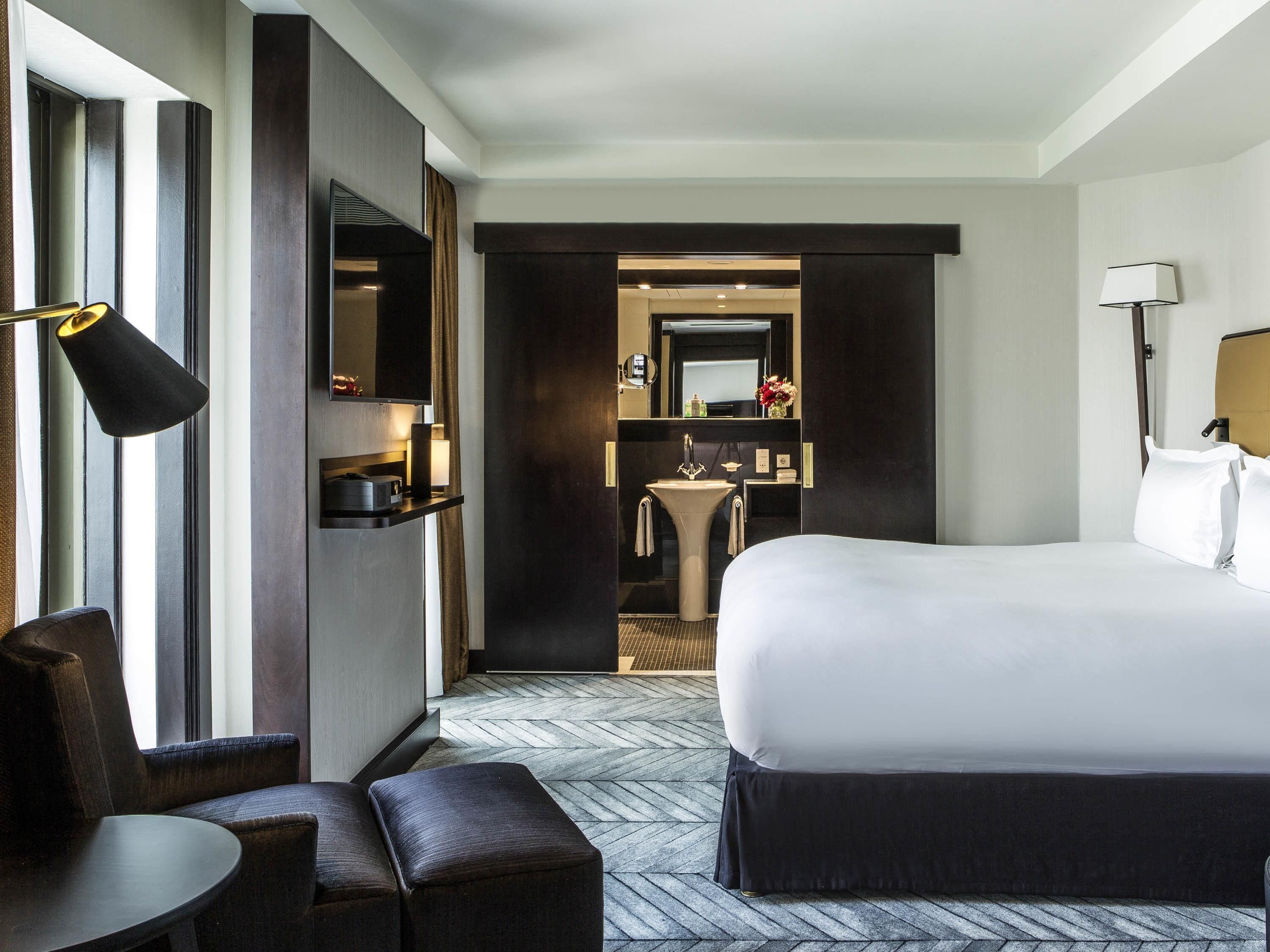A hotel room with a large bed, a desk, a chair, a sofa, a bathroom with a sink and mirror, and a TV.