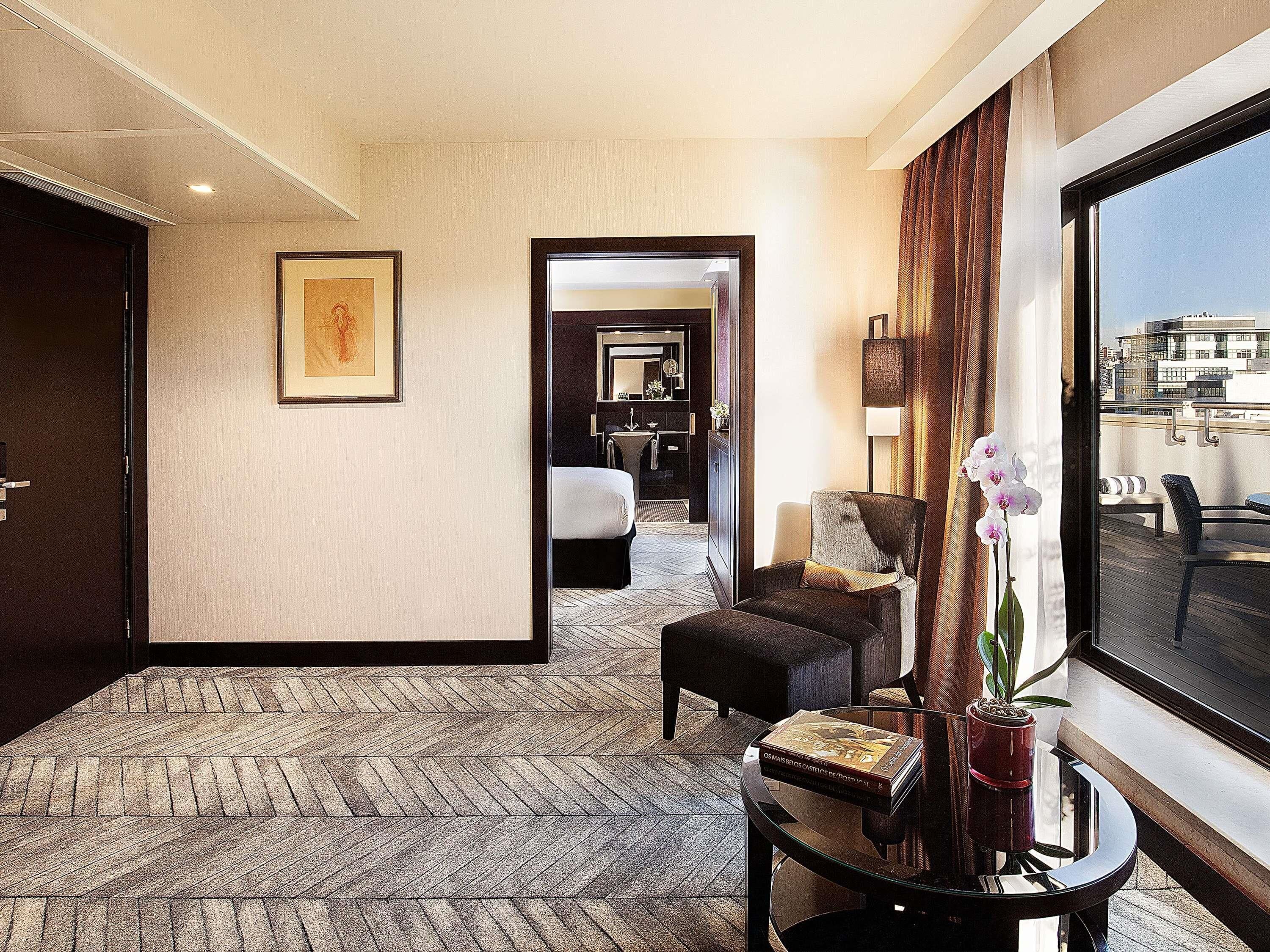 Habitación de hotel con alfombra de diseño espiga, un cuadro en la pared, una zona de estar con una silla y reposapiés, una mesita con un libro y una planta en maceta, y un ventanal con vistas a edificios.