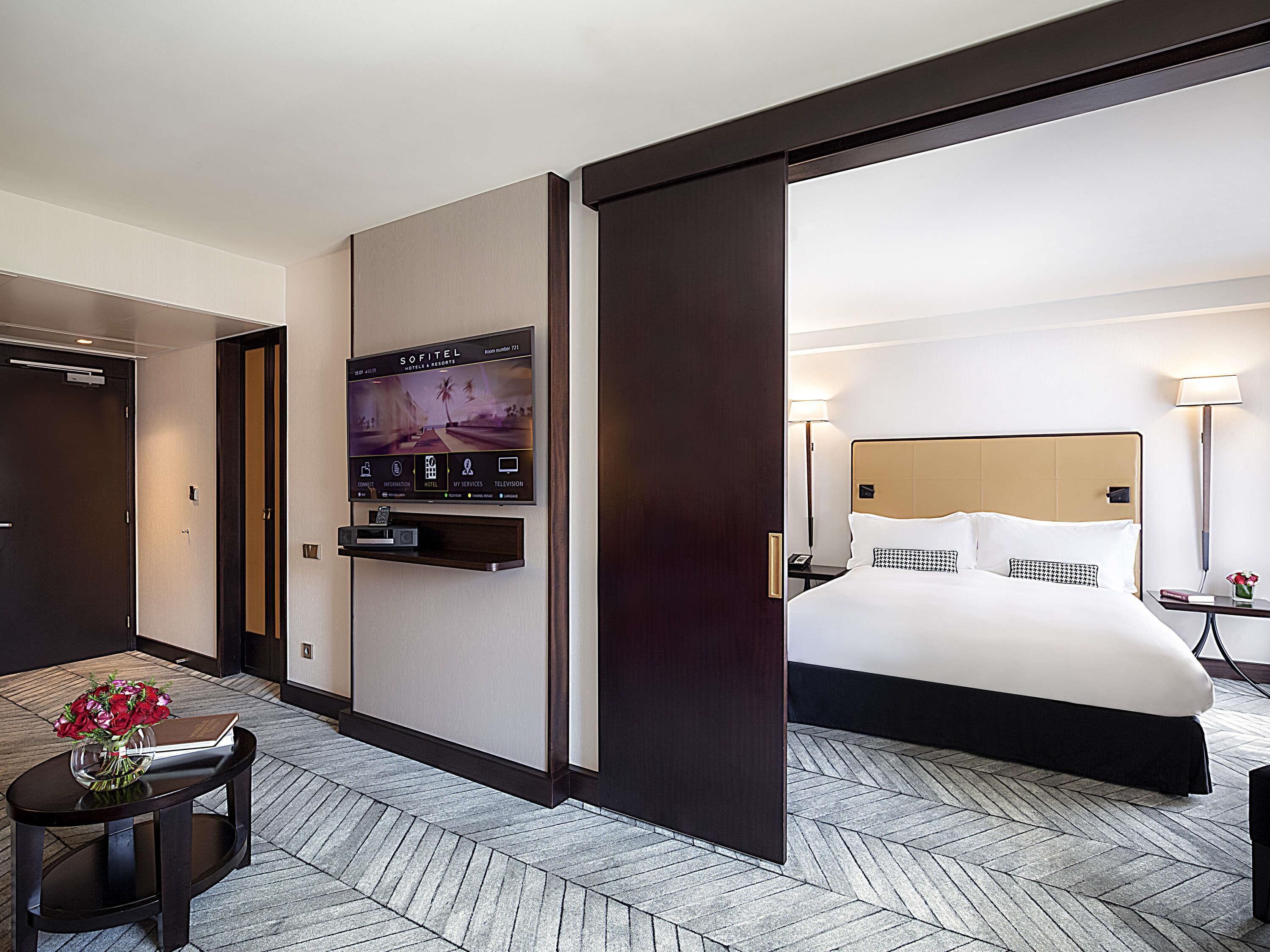 Habitación de hotel con cama, televisor montado en la pared, una mesita con un jarrón de flores y piso con estampado de espiga.