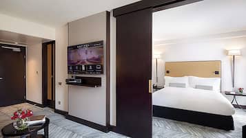 Habitación de hotel con cama, televisor montado en la pared, una mesita con un jarrón de flores y piso con estampado de espiga.