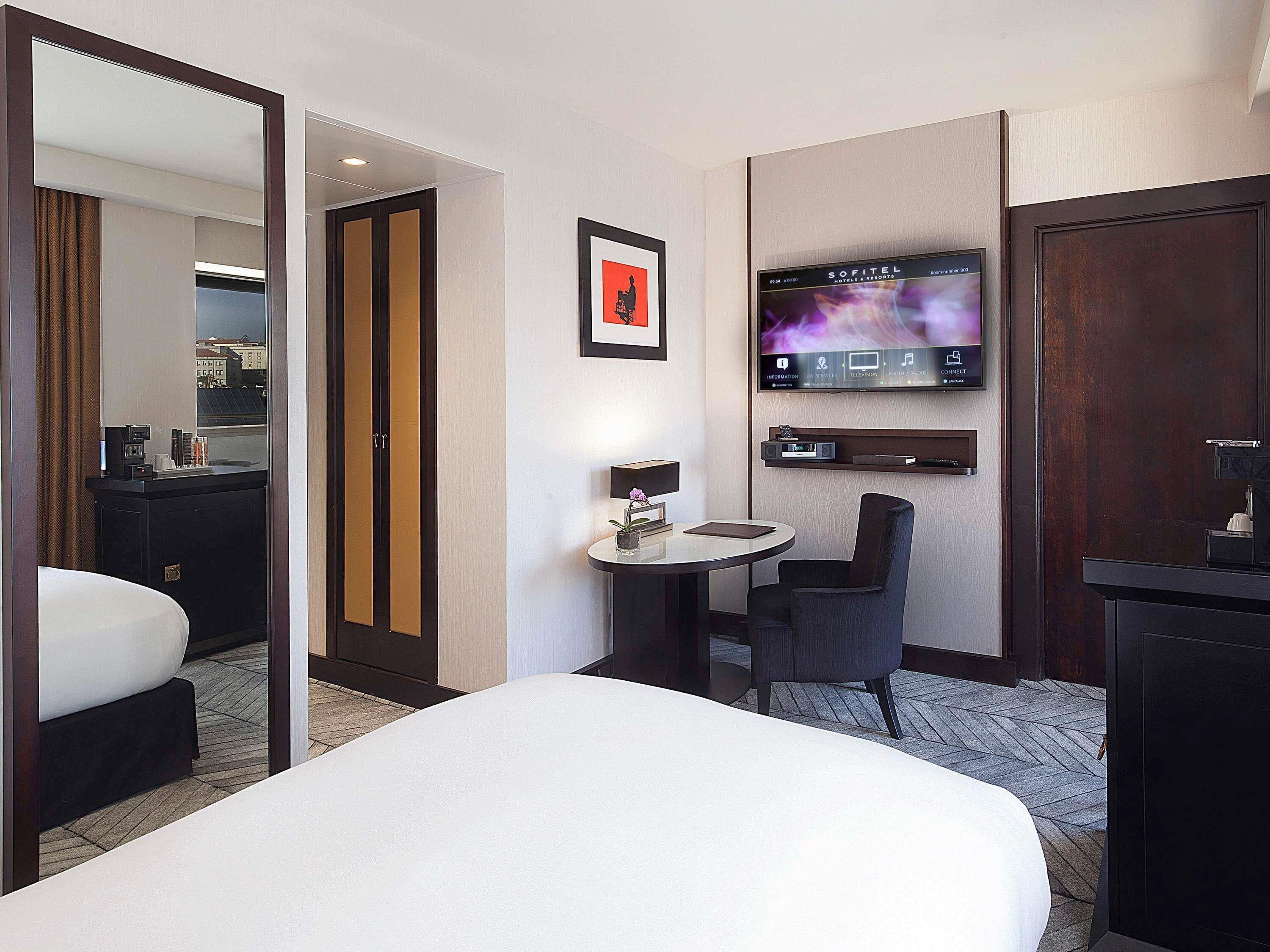 A hotel room with a bed, a desk with a chair, a TV mounted on the wall, and a minibar.