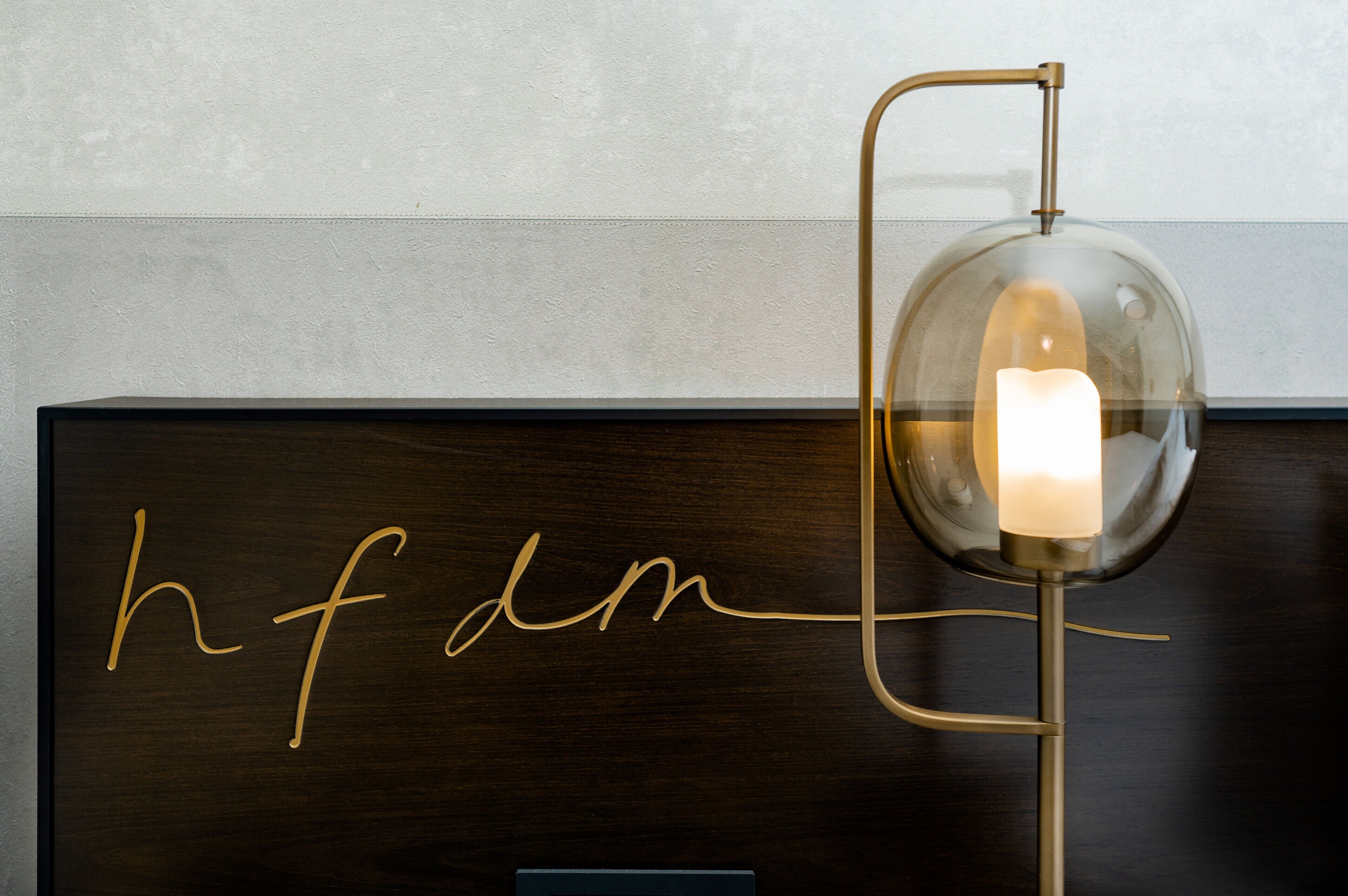 Une commode en bois foncé ornée d’un monogramme doré en lettres cursives, ainsi qu’une lampe de table moderne avec un abat-jour en verre transparent et une bougie à l’intérieur.