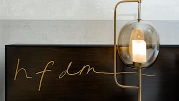 Une commode en bois foncé ornée d’un monogramme doré en lettres cursives, ainsi qu’une lampe de table moderne avec un abat-jour en verre transparent et une bougie à l’intérieur.
