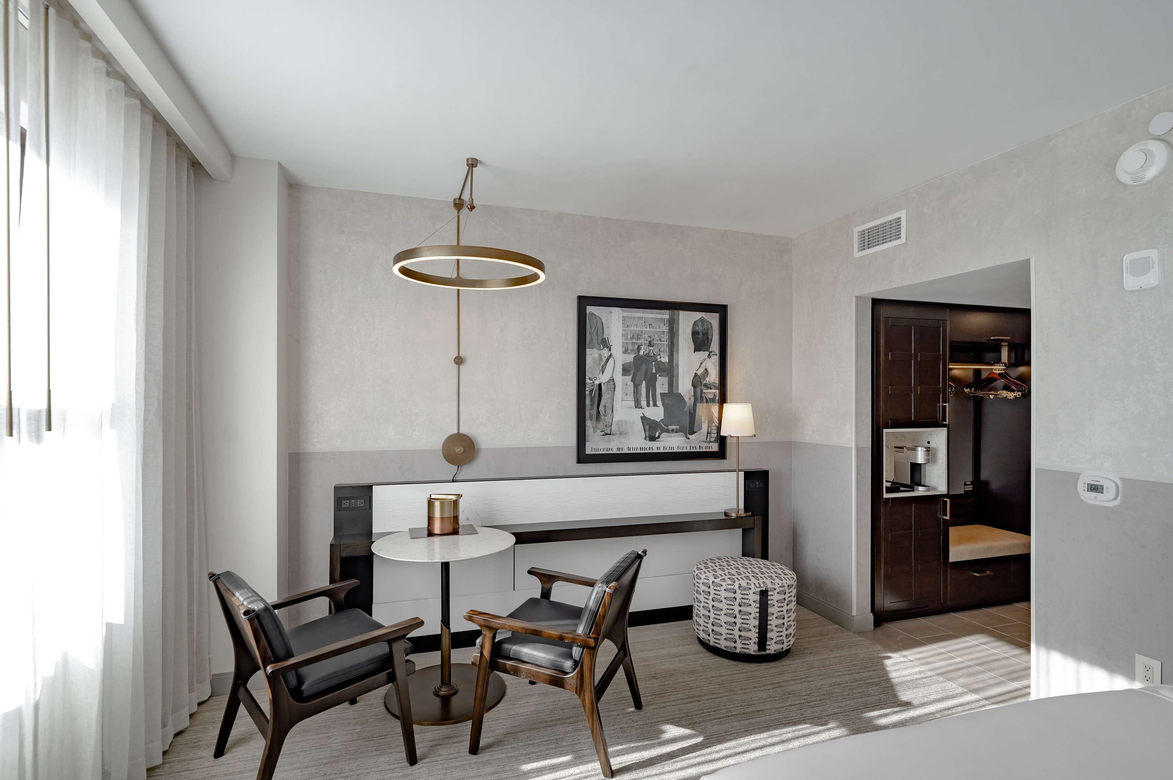 A modern hotel room with a round dining table, two chairs, a pendant light, a framed black and white photo, and a small round ottoman.