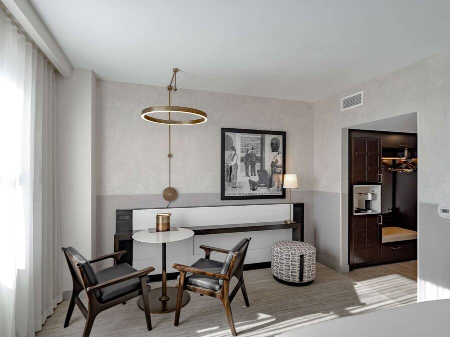 A modern hotel room with a round dining table, two chairs, a pendant light, a framed black and white photo, and a small round ottoman.