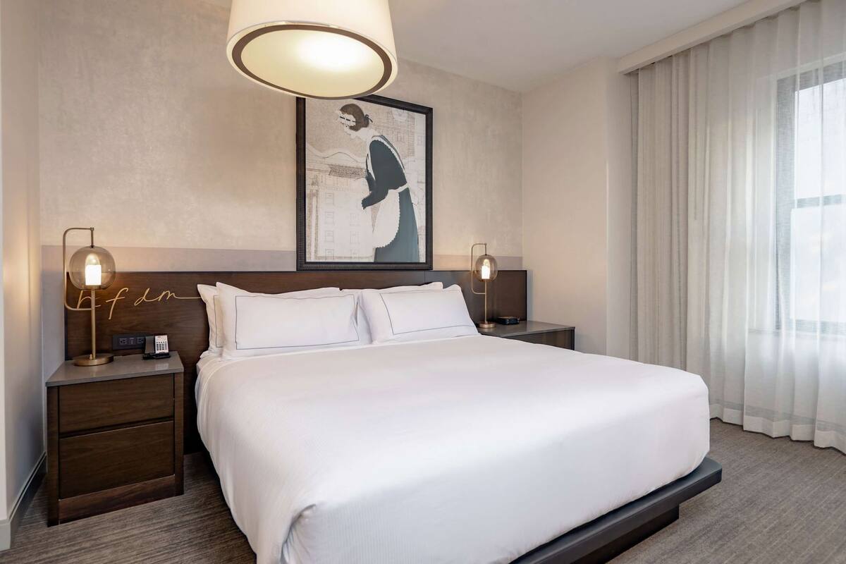 A hotel room with a large bed, two bedside tables, a framed artwork on the wall, and a window with curtains.
