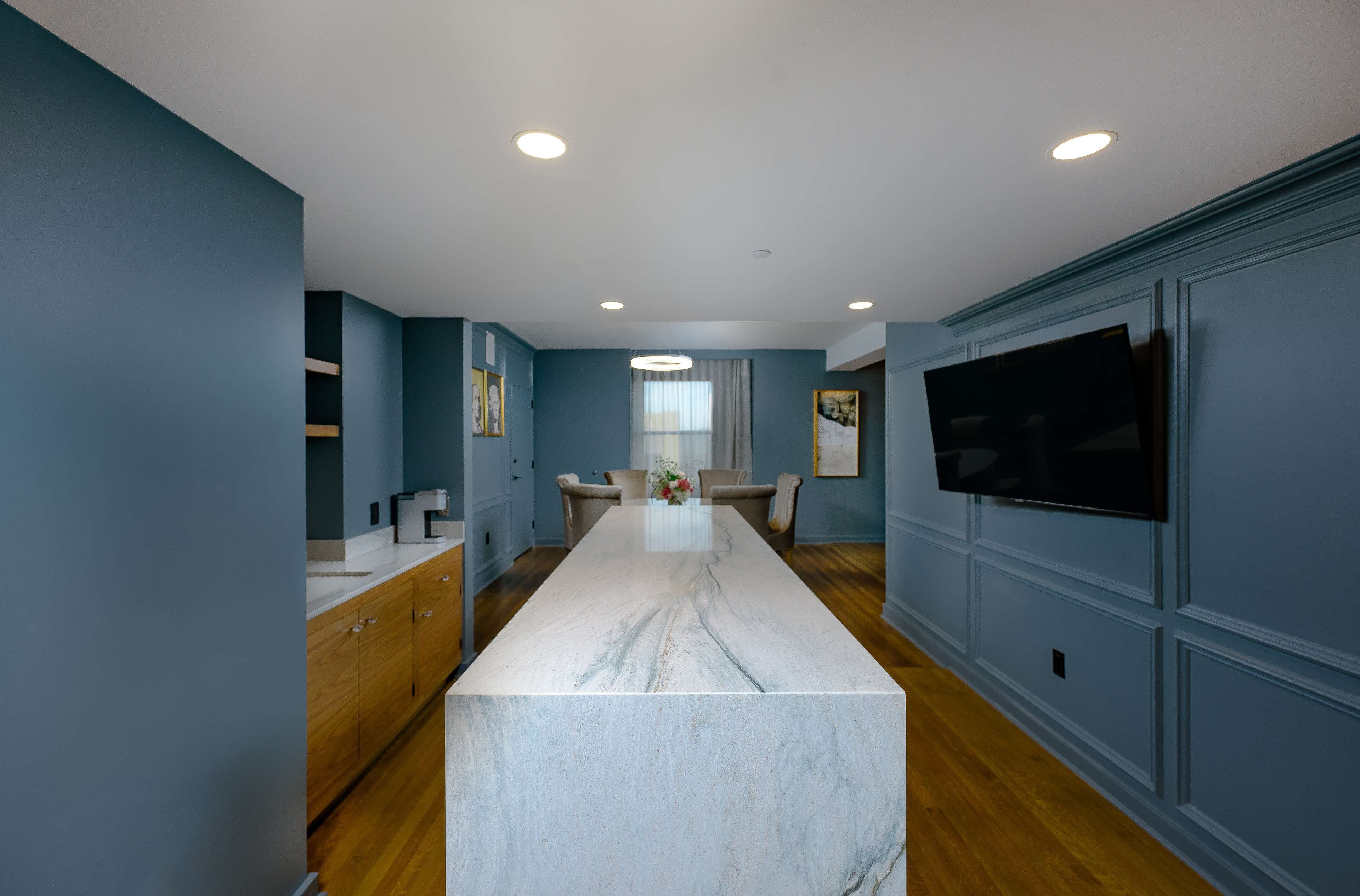 A modern kitchen with a marble island, wooden cabinets, and a flat-screen TV mounted on the wall.