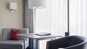 A hotel room with a gray sofa, a red patterned pillow, a small table with a book, a lamp, and a view through sheer curtains.