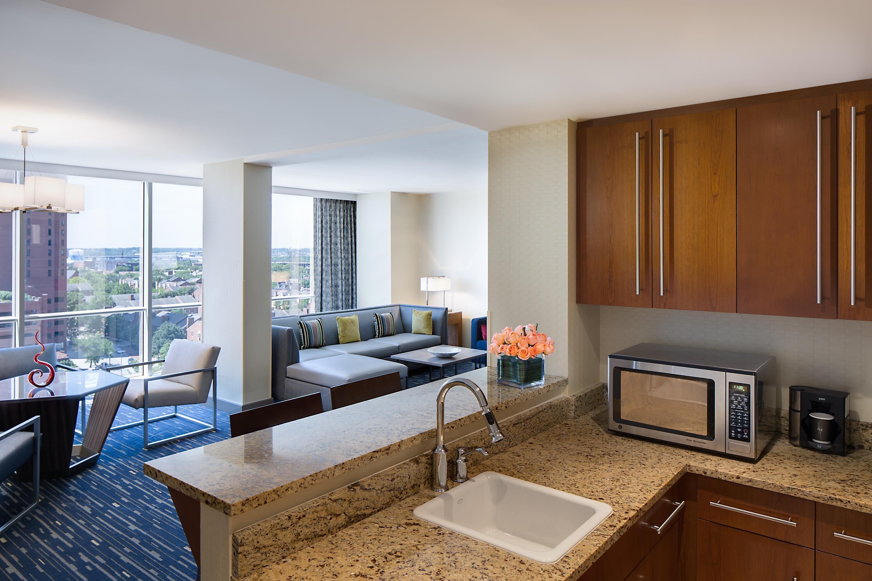 A modern hotel room with a kitchen area, a dining table, a sofa, and a view of the city.
