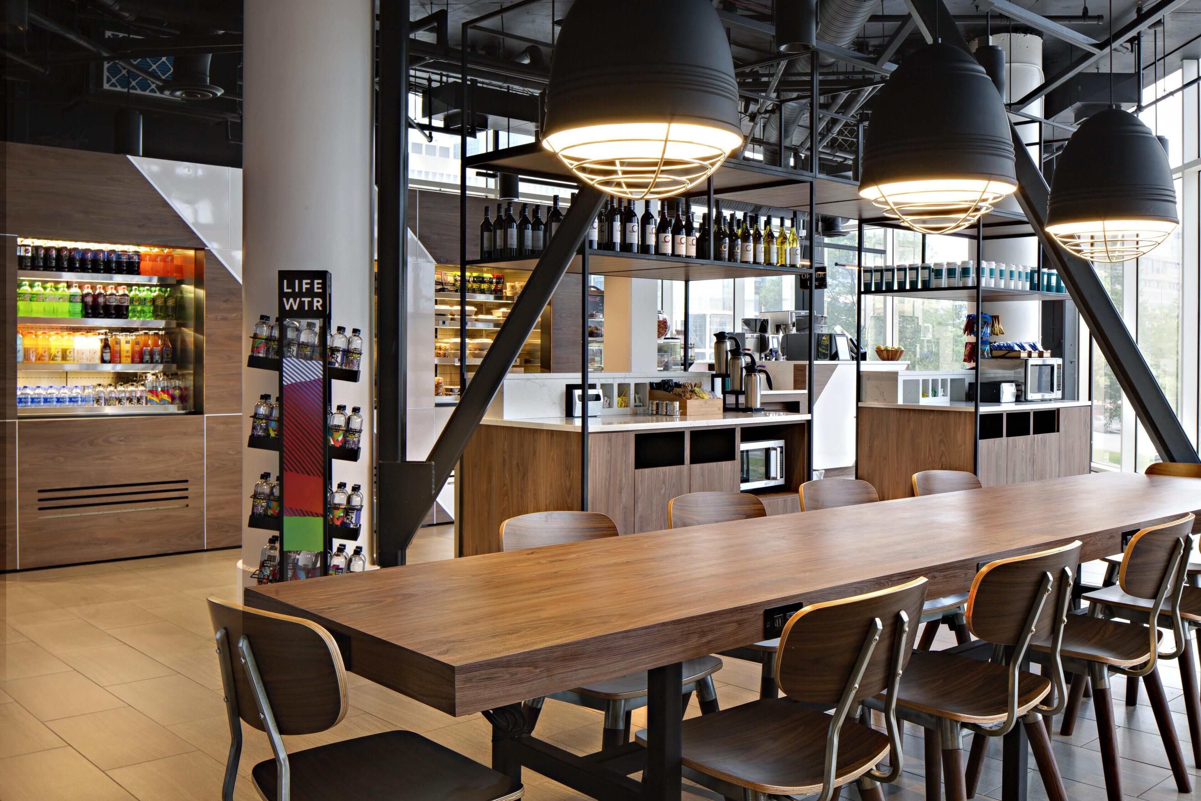 A modern restaurant interior with a long wooden table, multiple chairs, hanging pendant lights, a bar area with shelves of bottles, and a visible kitchen in the background.