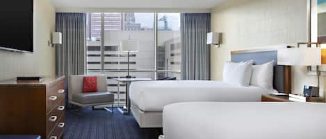 A hotel room with two beds, a desk, a chair, a TV, and a view of the city.