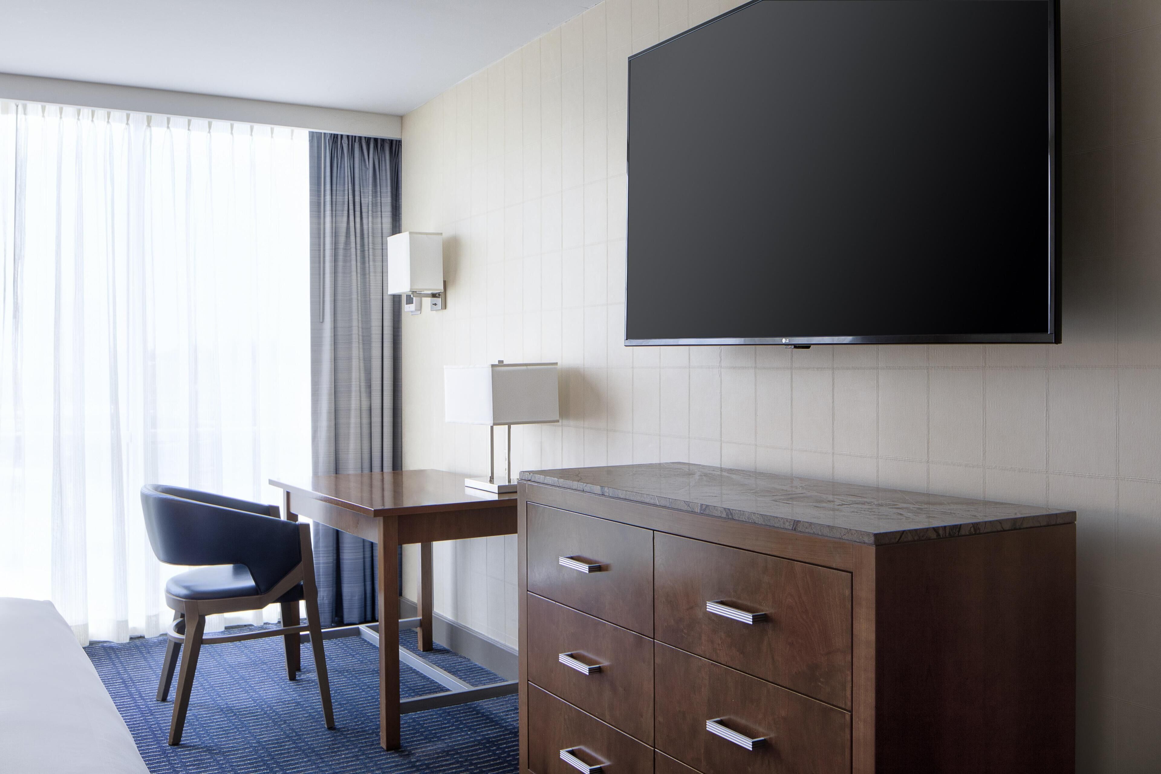 Une chambre d’hôtel comprenant un lit, un bureau avec une chaise, un téléviseur fixé au mur et une commode avec des tiroirs.