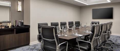 A conference room with a long table, multiple chairs, a large flat-screen TV, a mirror, and a minibar.