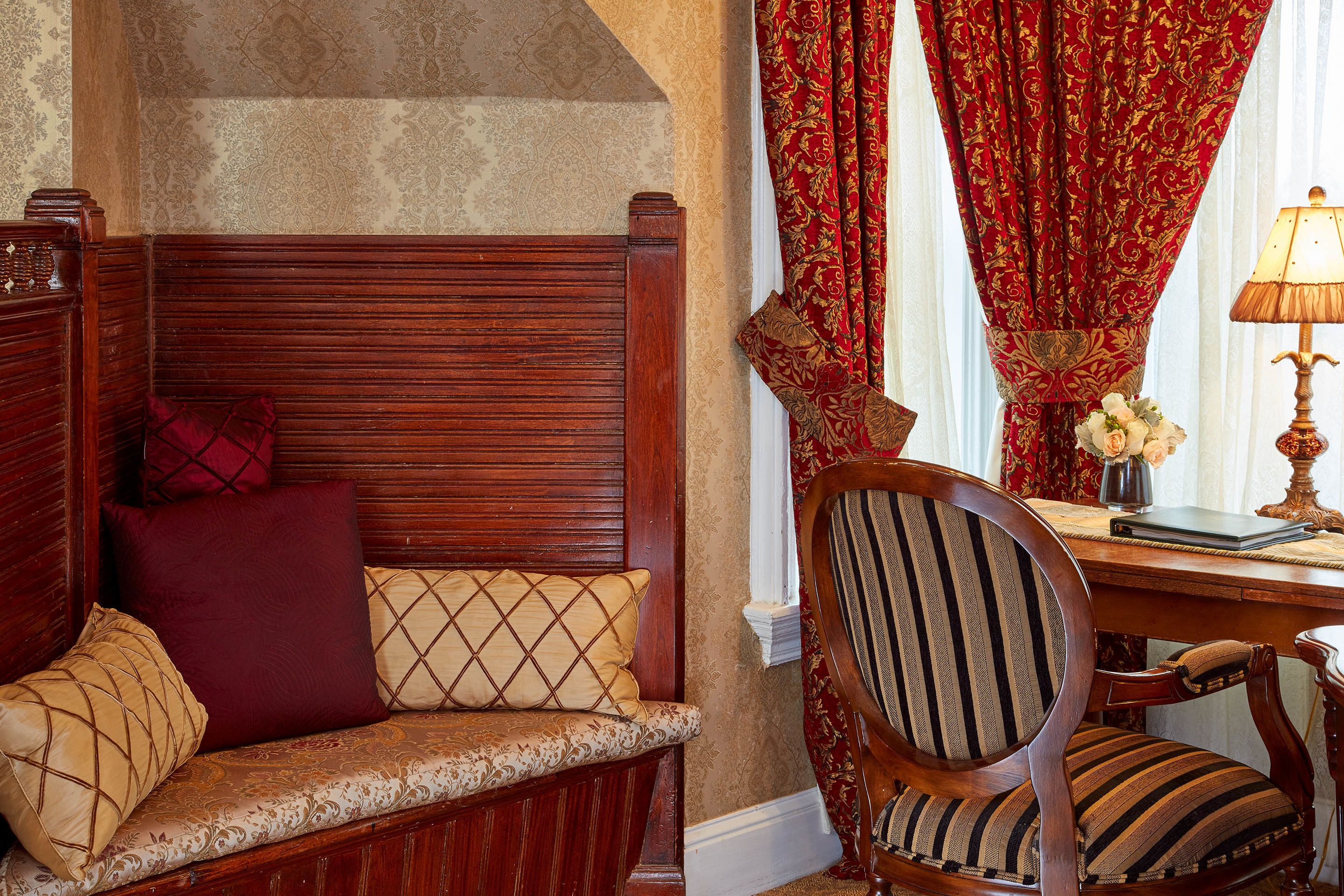 A wooden bench with patterned cushions, a striped upholstered chair, and a desk with a lamp.