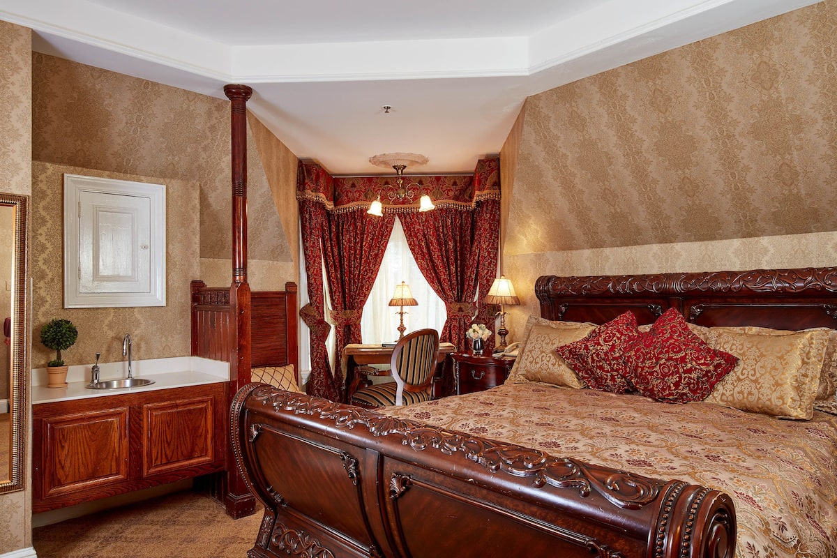 A bedroom with a large bed, a wooden headboard, a bedside table, a nightstand, a lamp, a chair, a desk, a mirror, and a window with curtains.