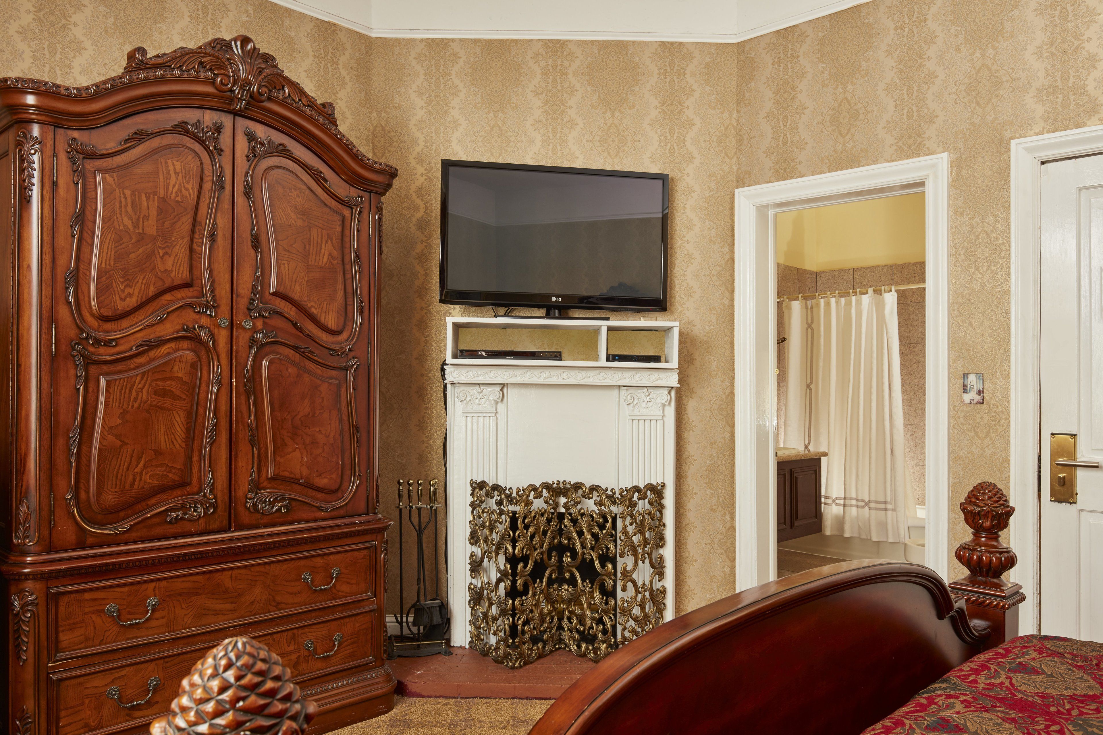 A bedroom with a wooden wardrobe, a television on a stand, a fireplace, and a door leading to a bathroom.