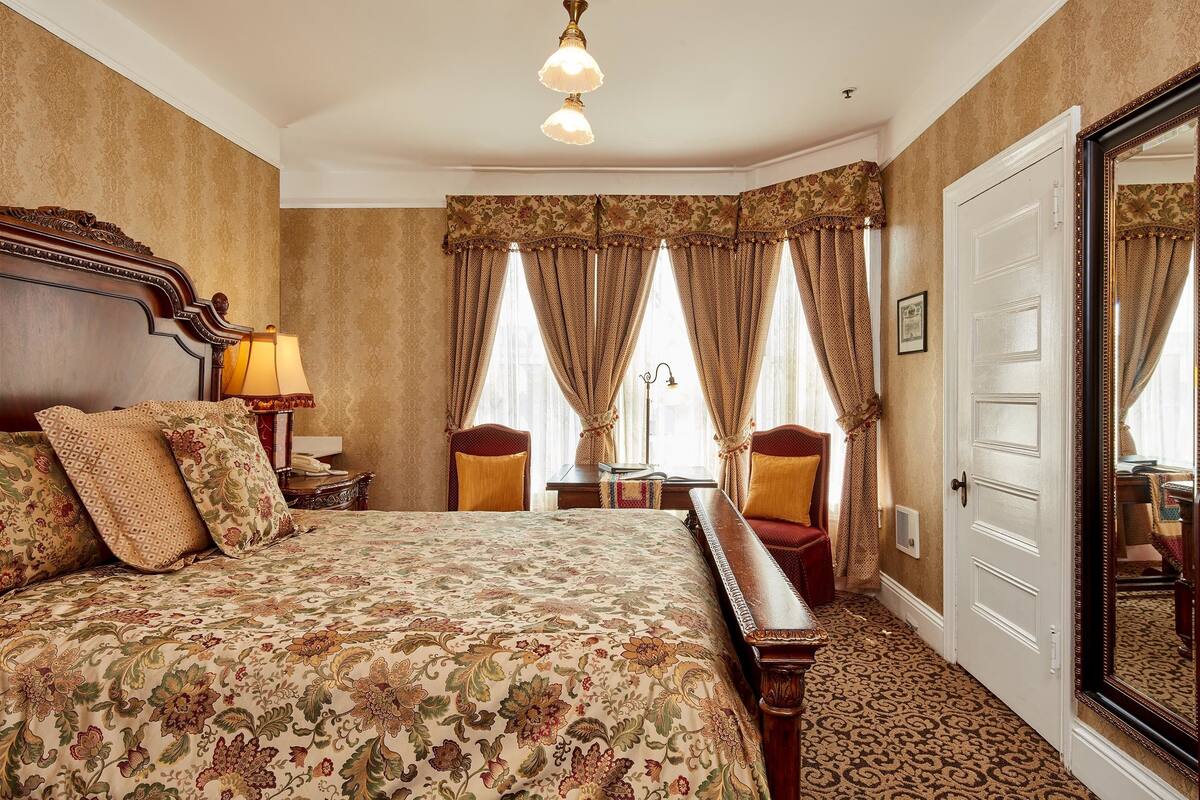 A bedroom with a large bed, two chairs, a nightstand, a lamp, and a door with a mirror.