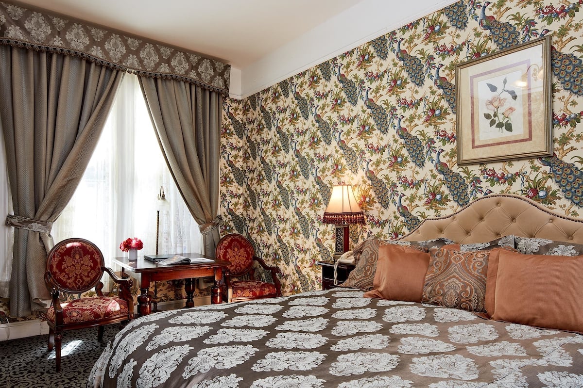 A bedroom with a large bed, a desk, two chairs, and a wall with floral wallpaper.