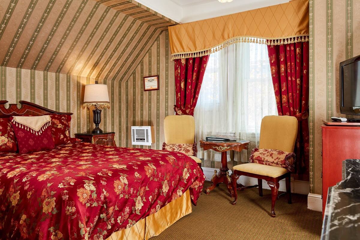 A hotel room with a large bed, two chairs, a small table, a TV, and a window with curtains.