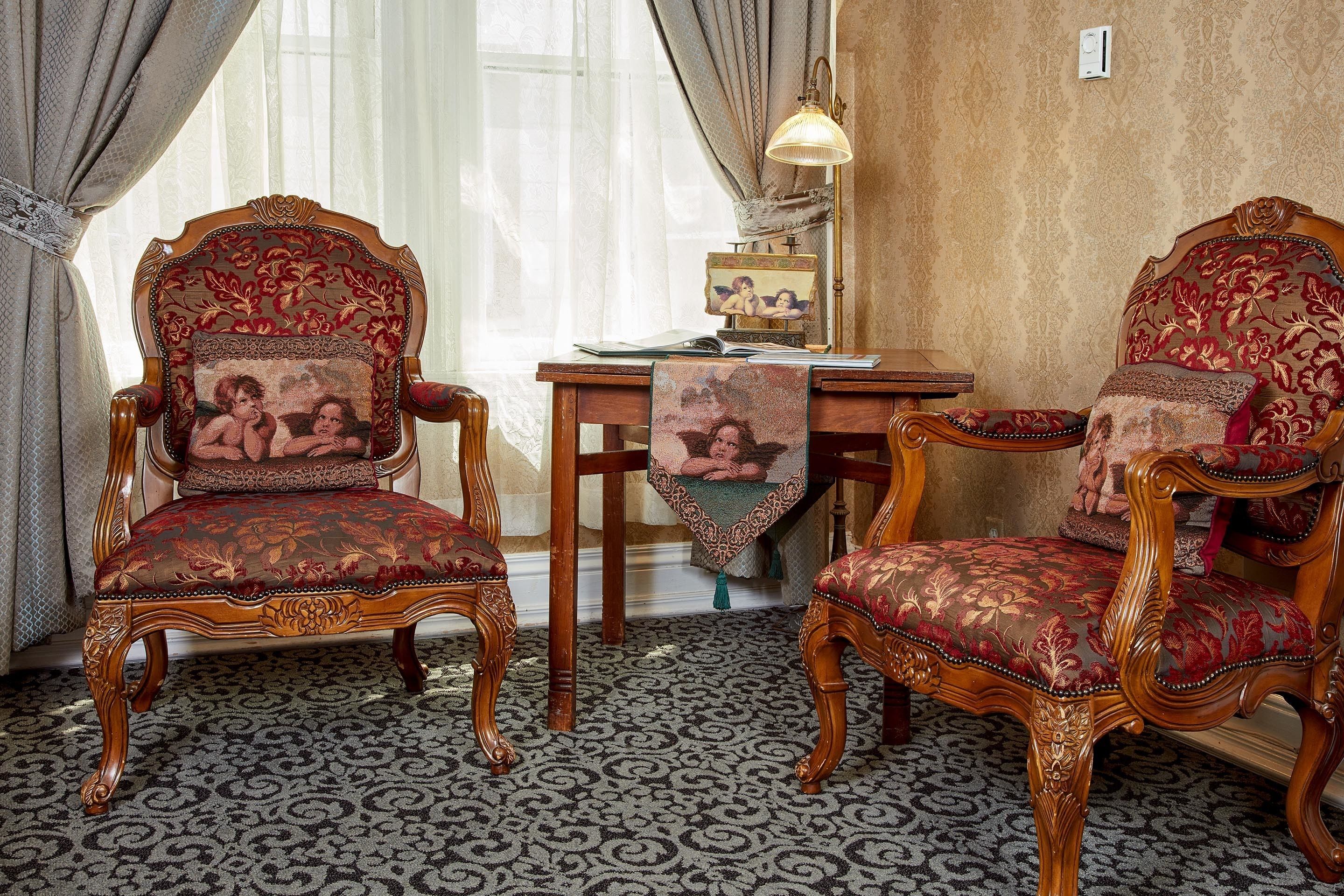 Two ornate chairs with floral upholstery, a patterned carpet, and a table with a lamp and picture frame.