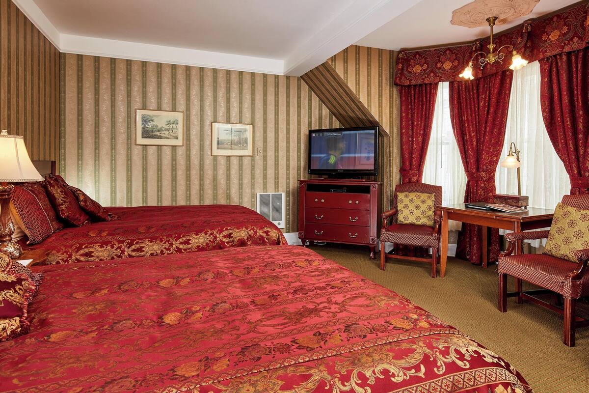 A hotel room with two large beds, a TV, a desk with chairs, and a chandelier.