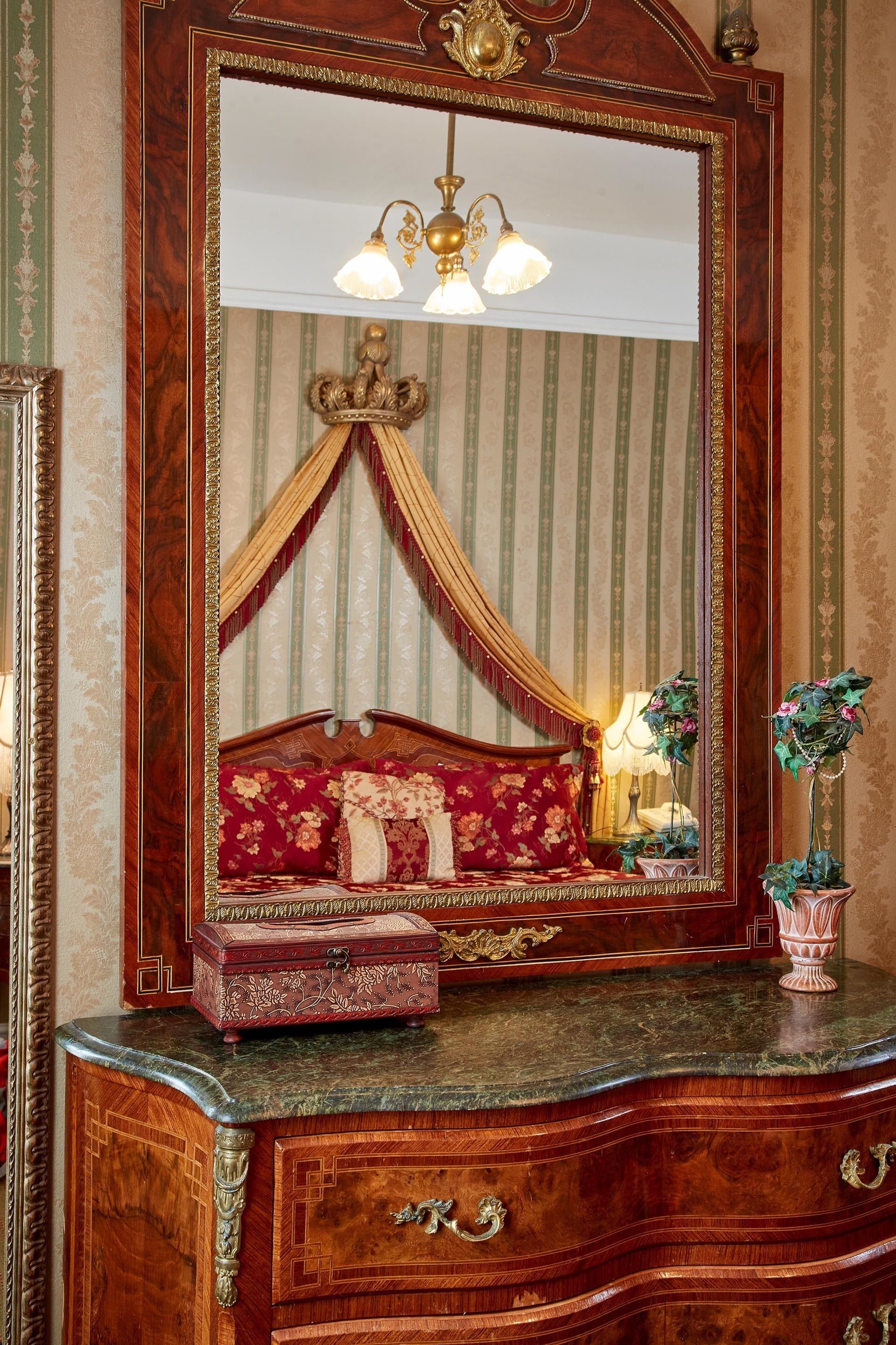 A bedroom with a large ornate mirror, a canopy bed, a dresser with drawers, and a vase with flowers.