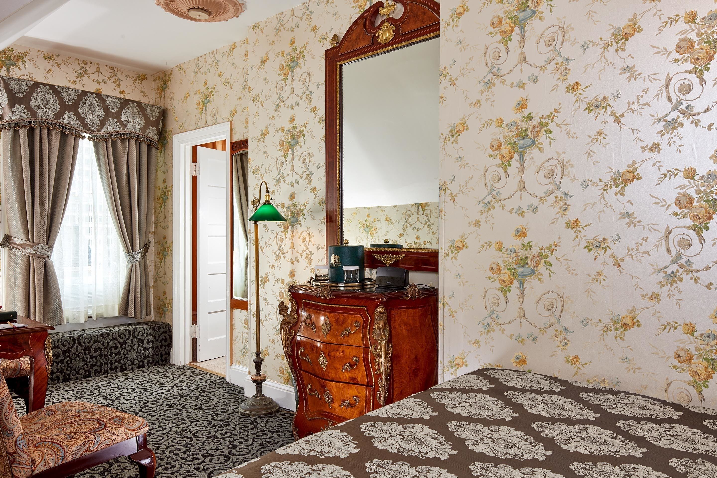A room with floral wallpaper, a wooden dresser, a bench, and a bed with patterned bedding.