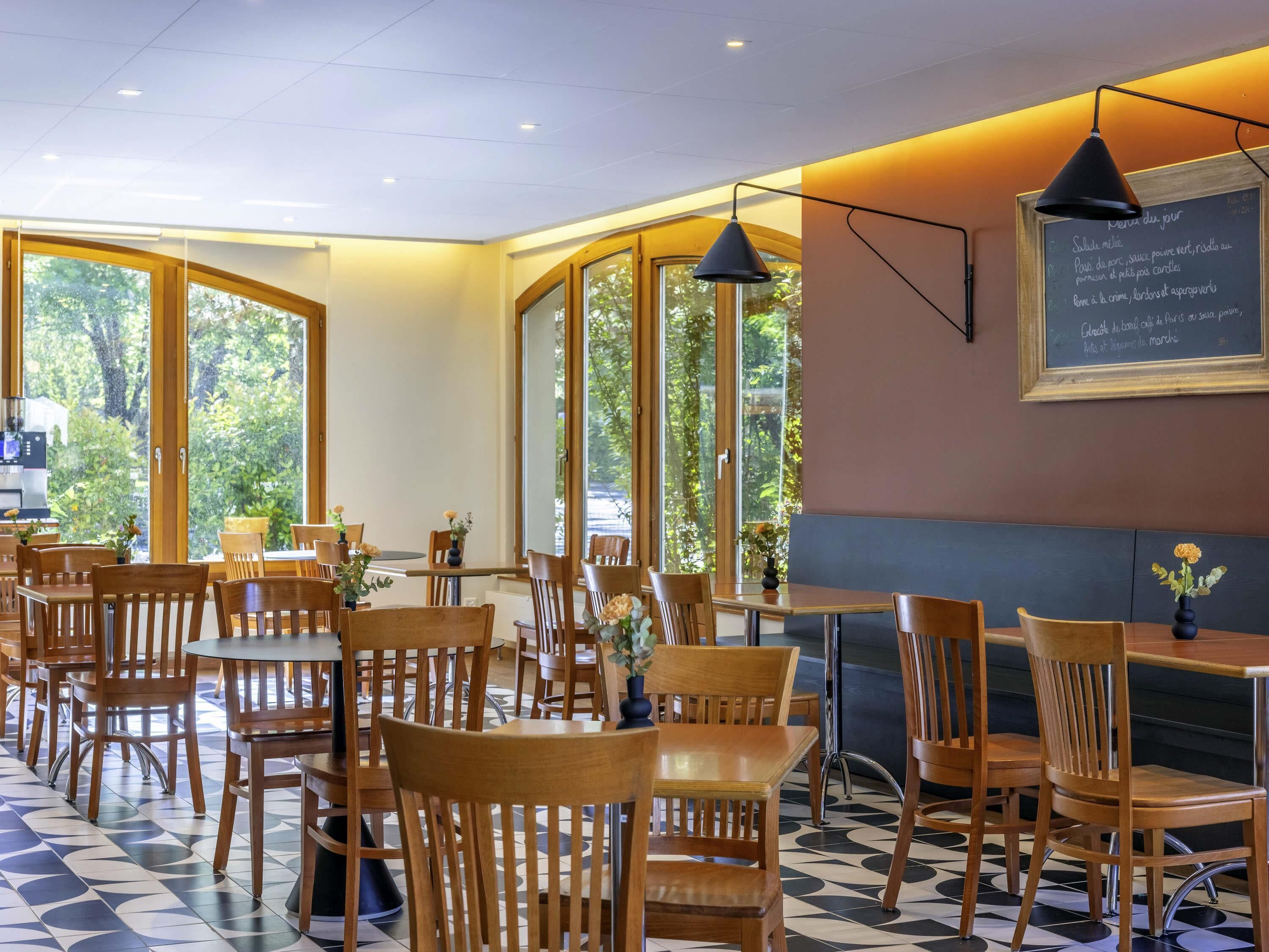 A restaurant with wooden chairs and tables, large windows, and a chalkboard menu.